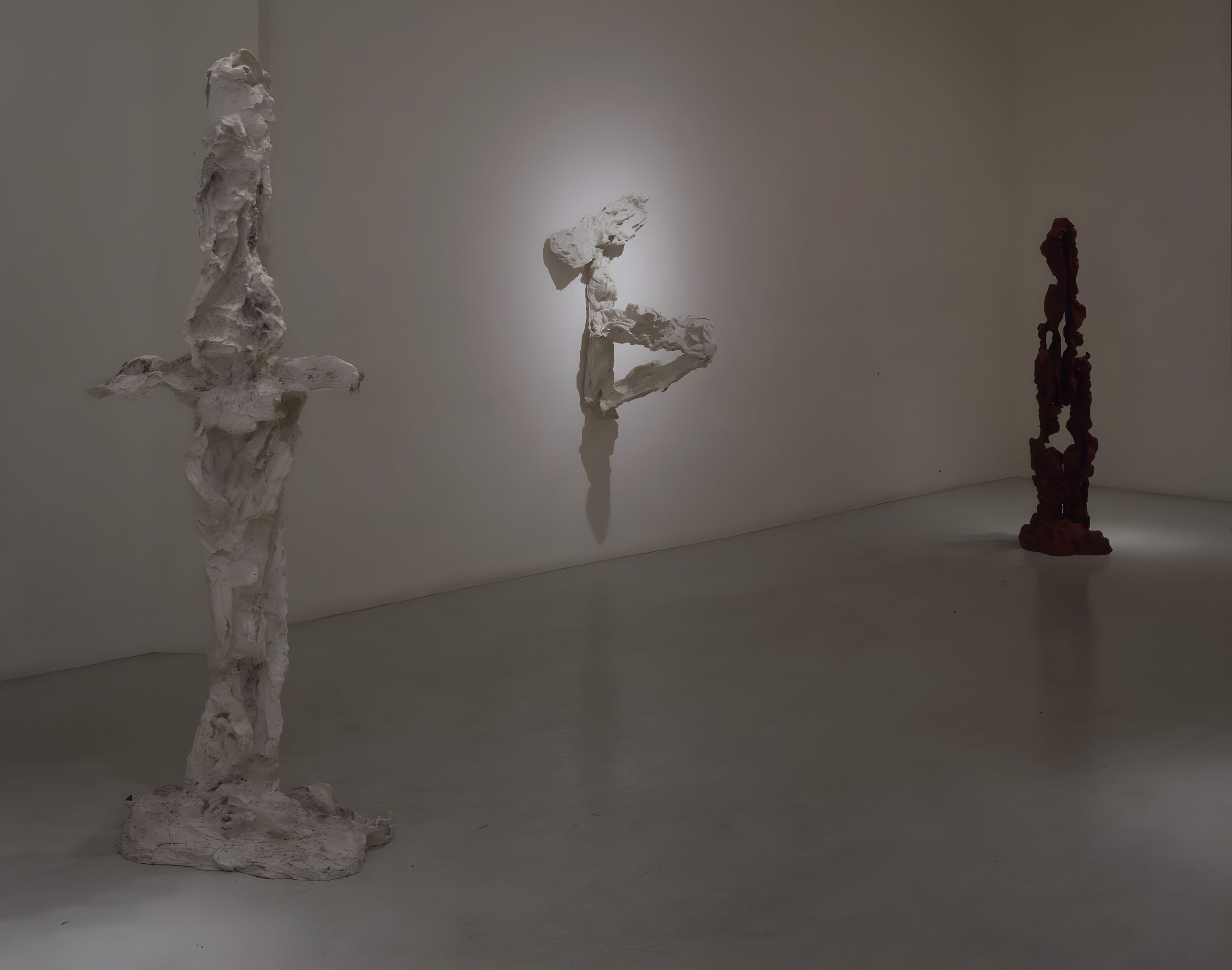 Alain Kirili, Exposition, installation view, 1994: three standing sculptures, one tall and pale, one small on the wall and one dark red, in a gray gallery.
