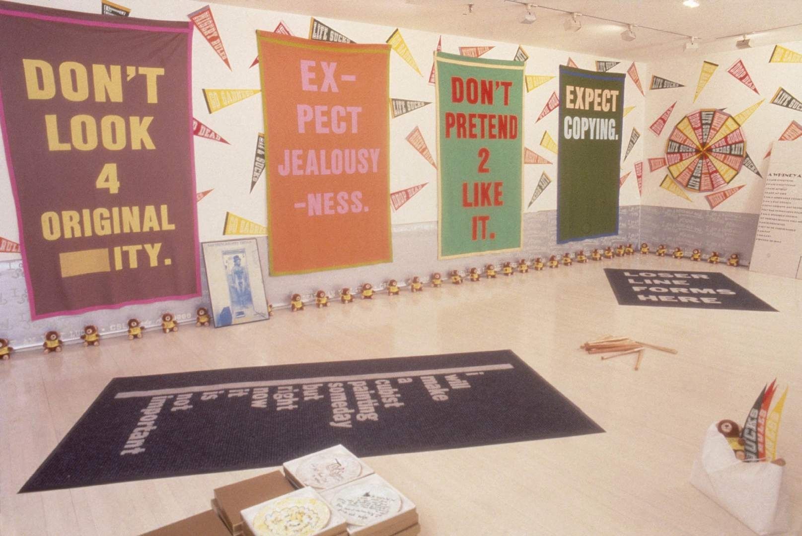 Cary Leibowitz (Candyass), installation view, 1989: brightly coloured text banners and pennants on the gallery walls, with a printed rug on the floor.