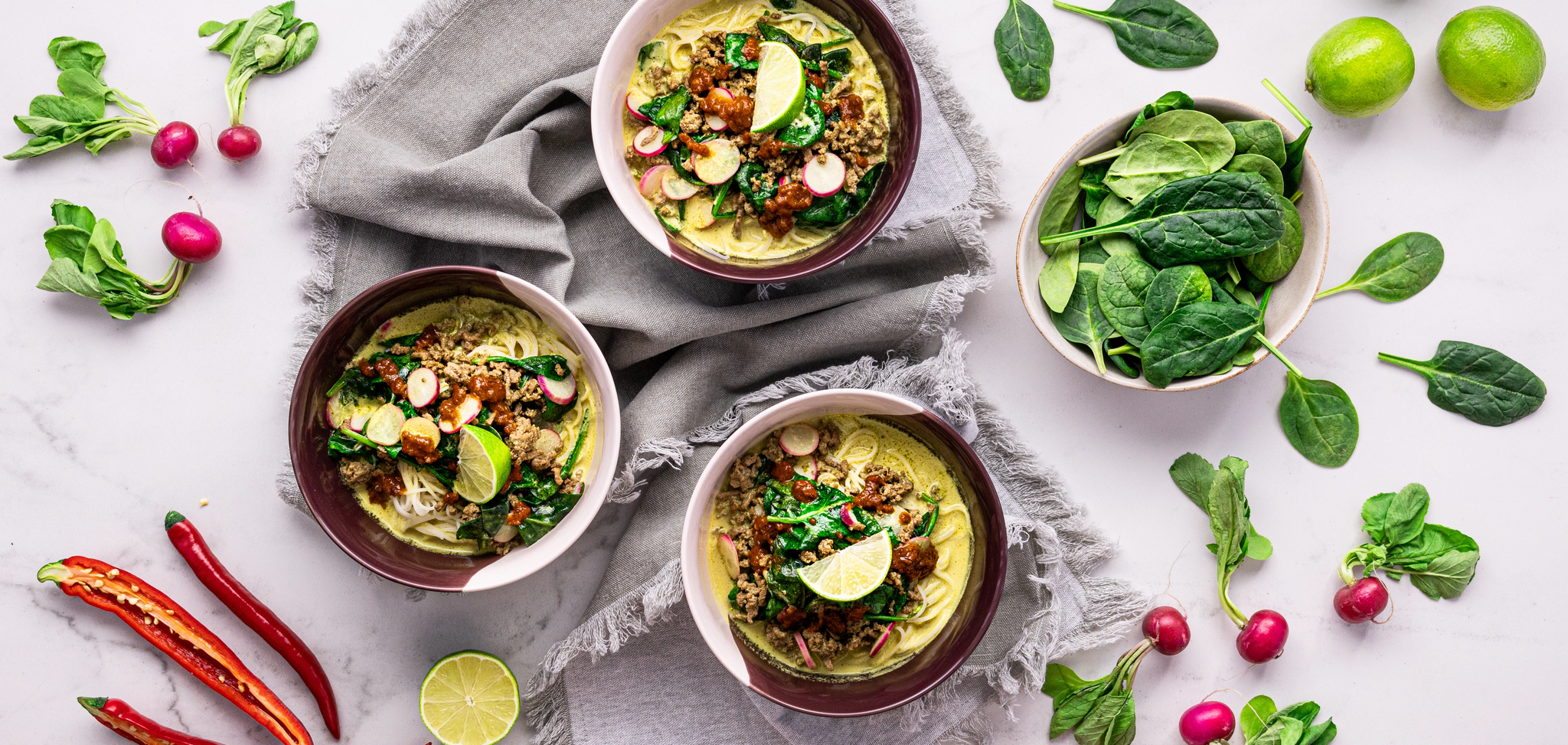 Three bowls of creamy coconut soup on a table with fresh spinach, limes and chillies beside them
