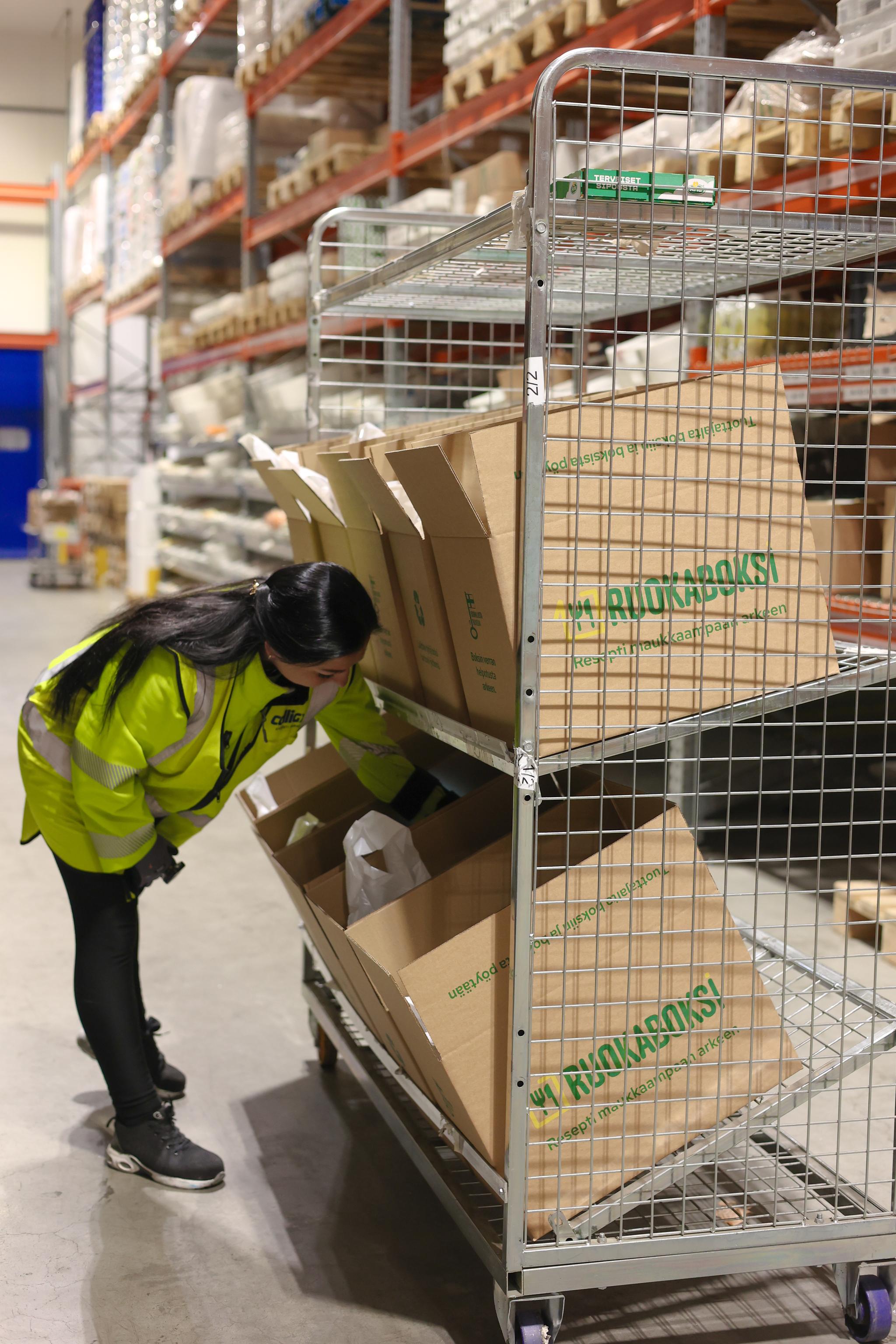 A worker in a yellow vest examines meal kit boxes on a rolling cart in a warehouse.