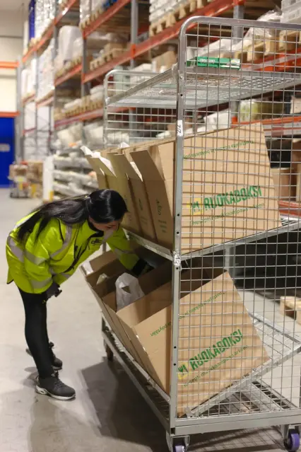 A worker in a yellow vest examines meal kit boxes on a rolling cart in a warehouse.