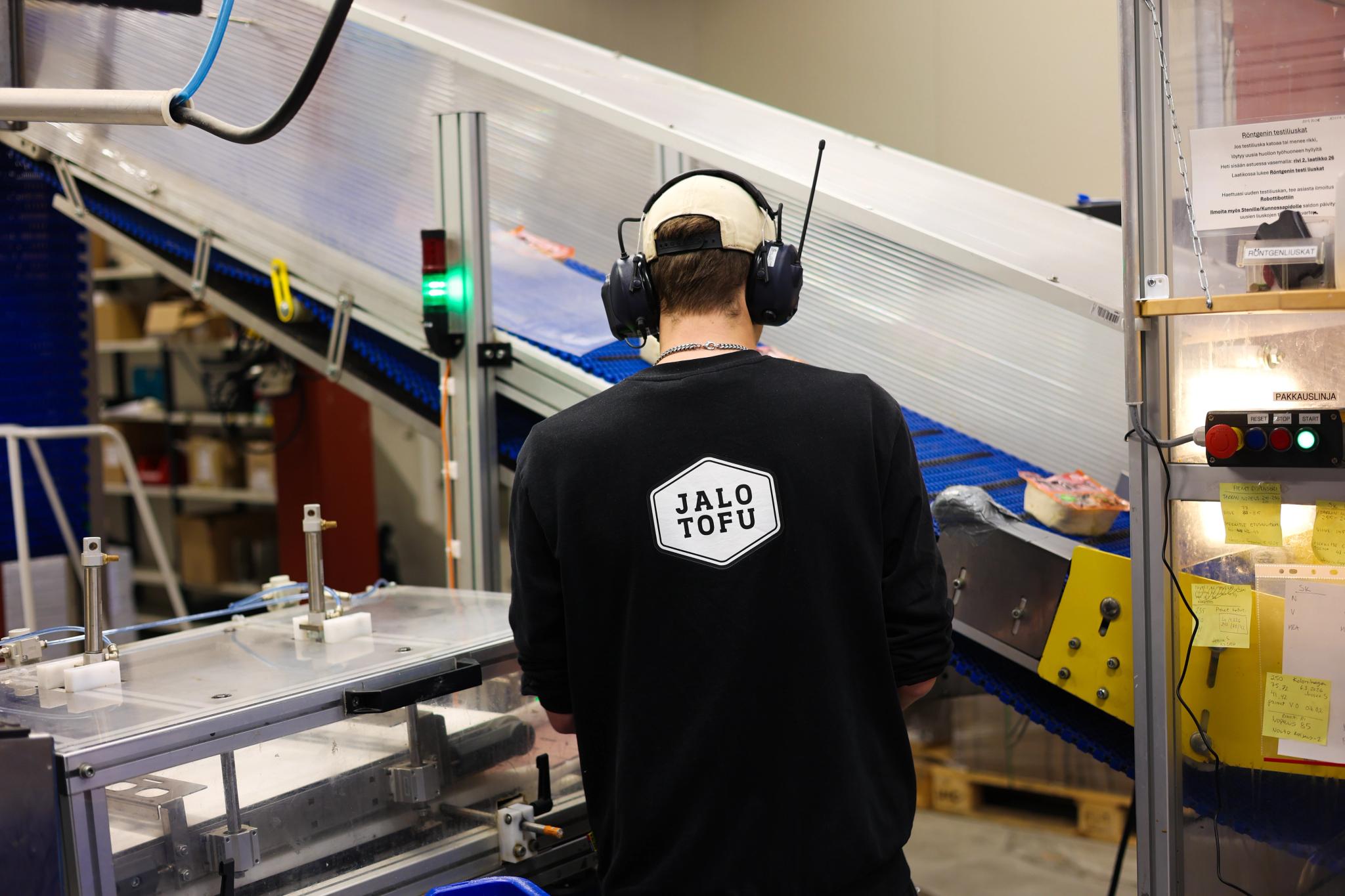 A person wearing a JALO TOFU shirt and ear protection observes a factory production line.