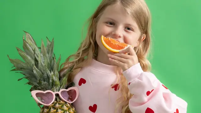 Tyttö hymyilee appelsiini suussa ja ananas sylissään