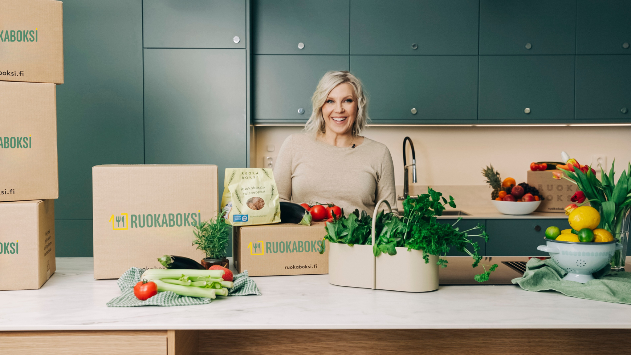 A smiling woman in a modern kitchen with several RUOKABOKSI meal kit boxes and abundant fresh produce.