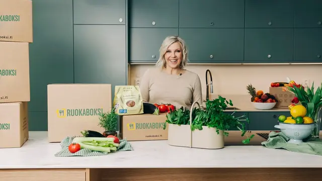 A smiling woman in a modern kitchen with several RUOKABOKSI meal kit boxes and abundant fresh produce.