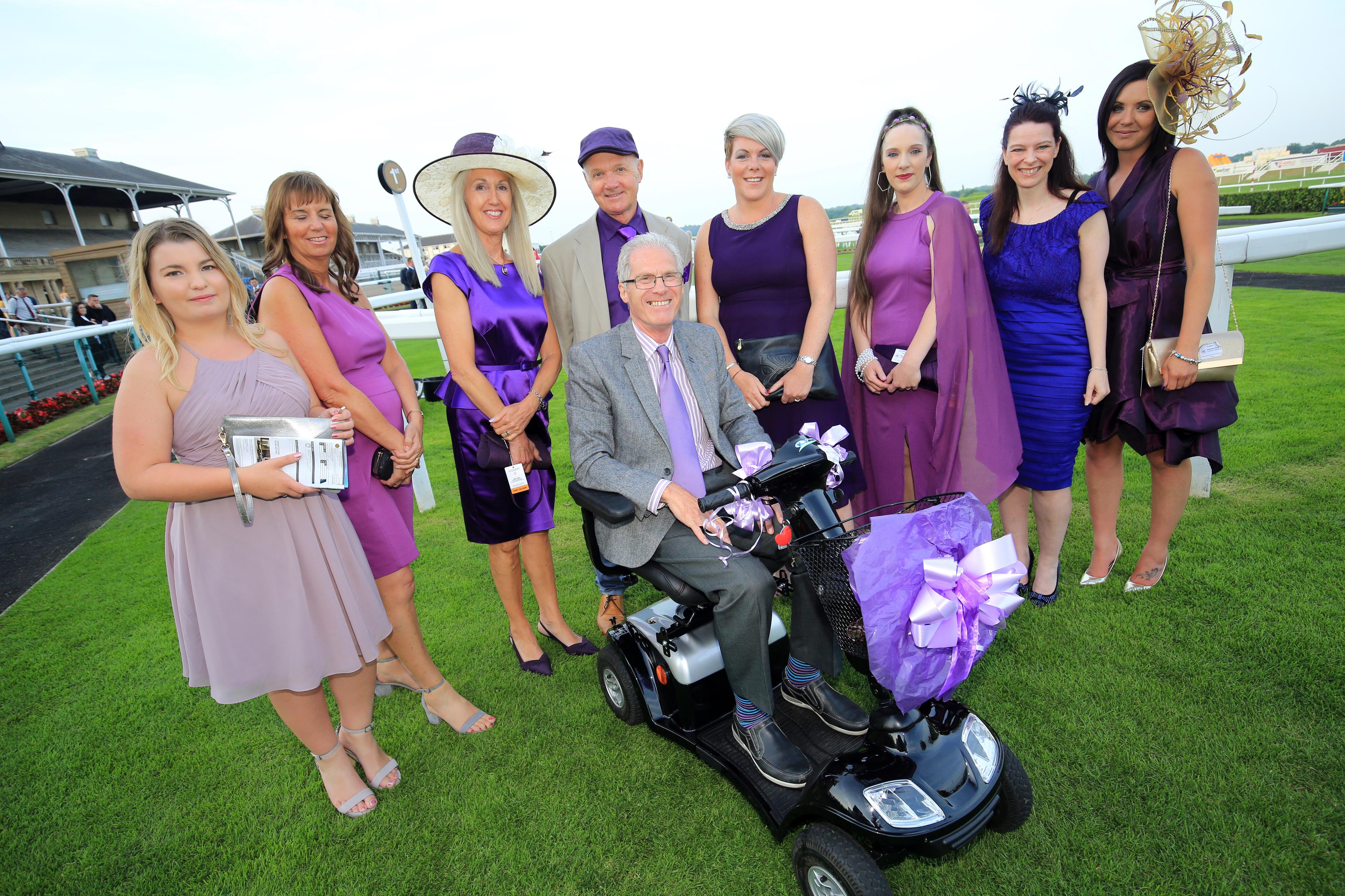 A group of people - seven women and one man - stand facing the camera, with another man sitting on a mobility scooter in the foreground. All those pictured are well-dressed, and are pictured outside at the Weston Park Cancer Charity Race Evening at Doncaster Racecourse.
