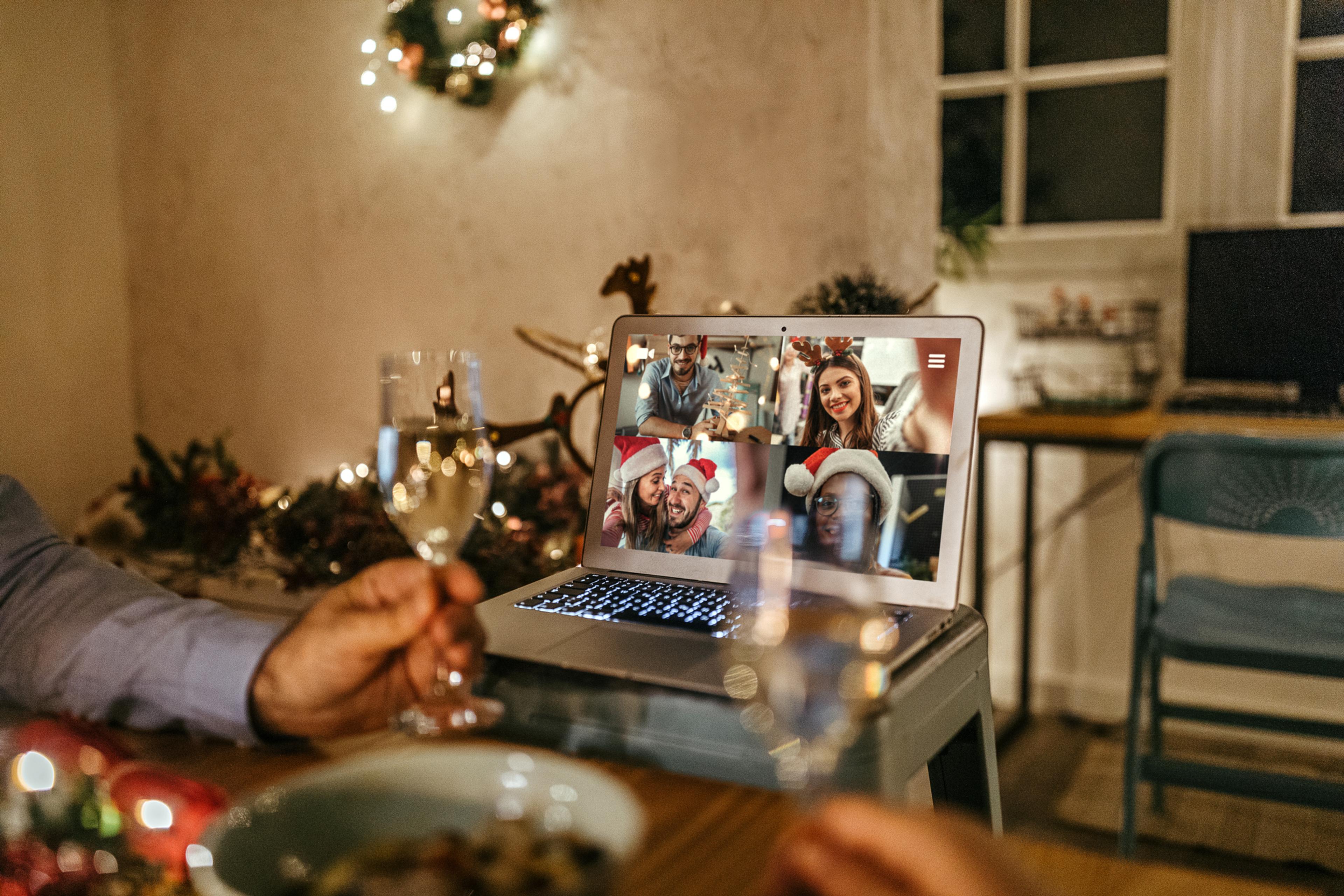 A laptop is resting on a chair with a video call on the screen, the setting is festive with fairy lights and a man in the foreground holding a glass of prosecco.