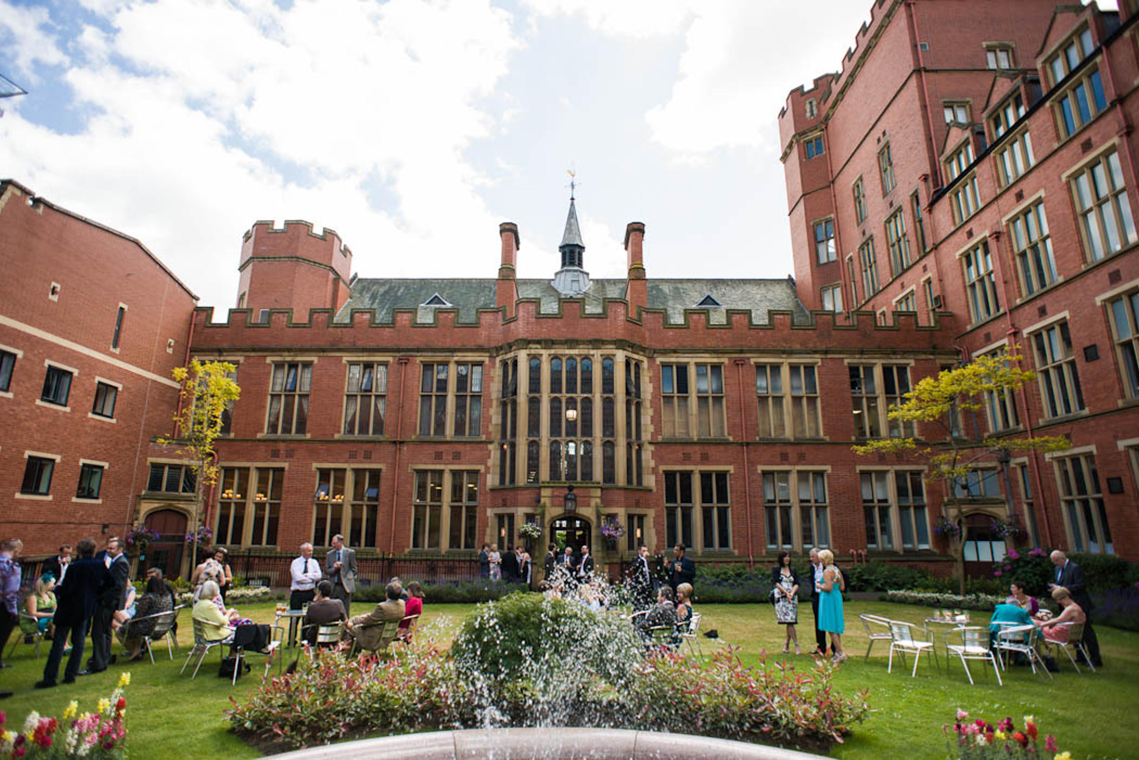 A courtyard featuring a fountain and a lawn, and formally-dressed people in front of a red brick building. The building is the University of Sheffield's Firth Court.