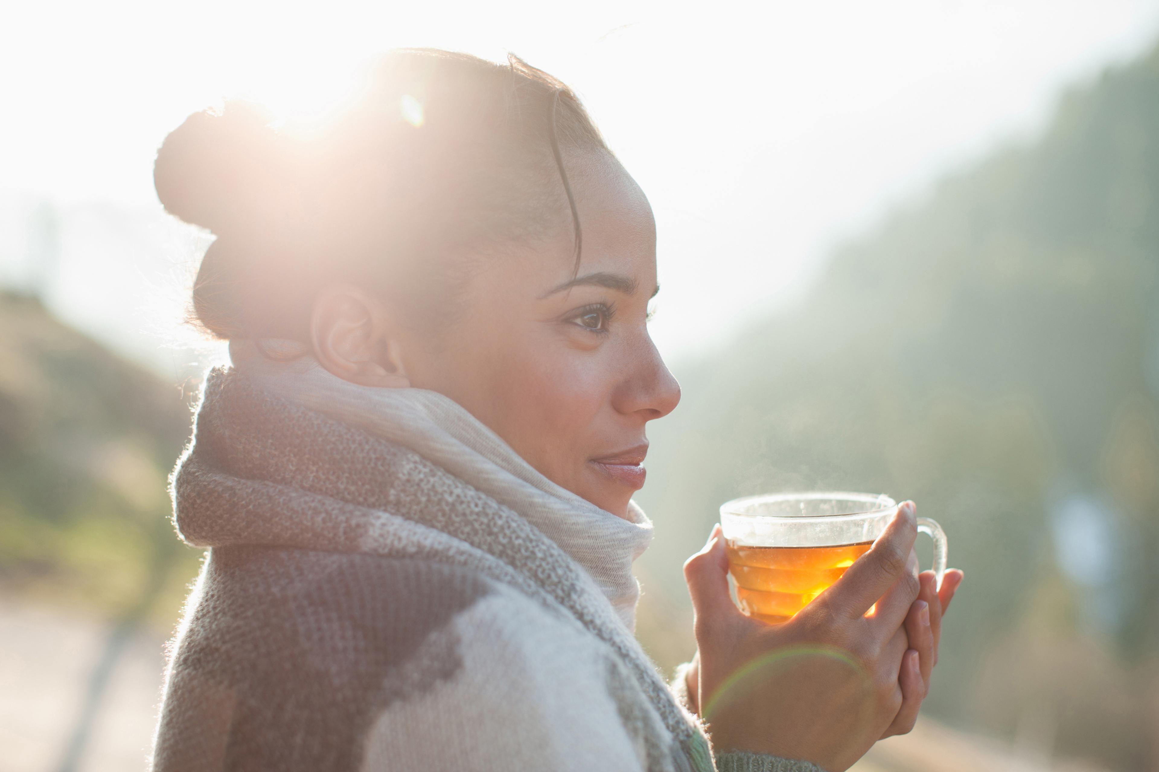 A side profile of a woman wrapped in a scarf and cardigan holding a glass cup of tea