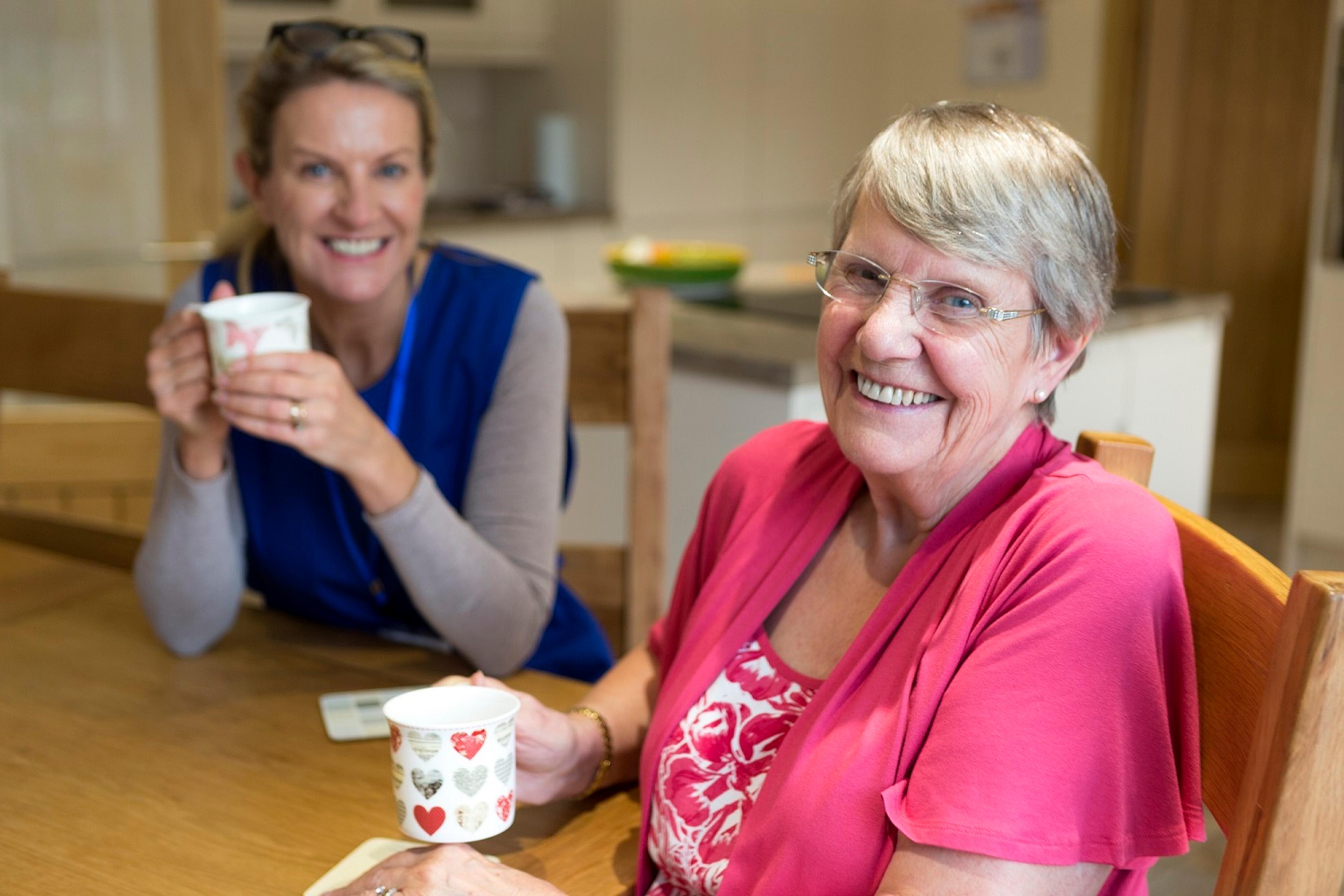 An older lady smiles at the camera with a cup of tea in her hand sat at a table, whilst a support worker sits next to her.
