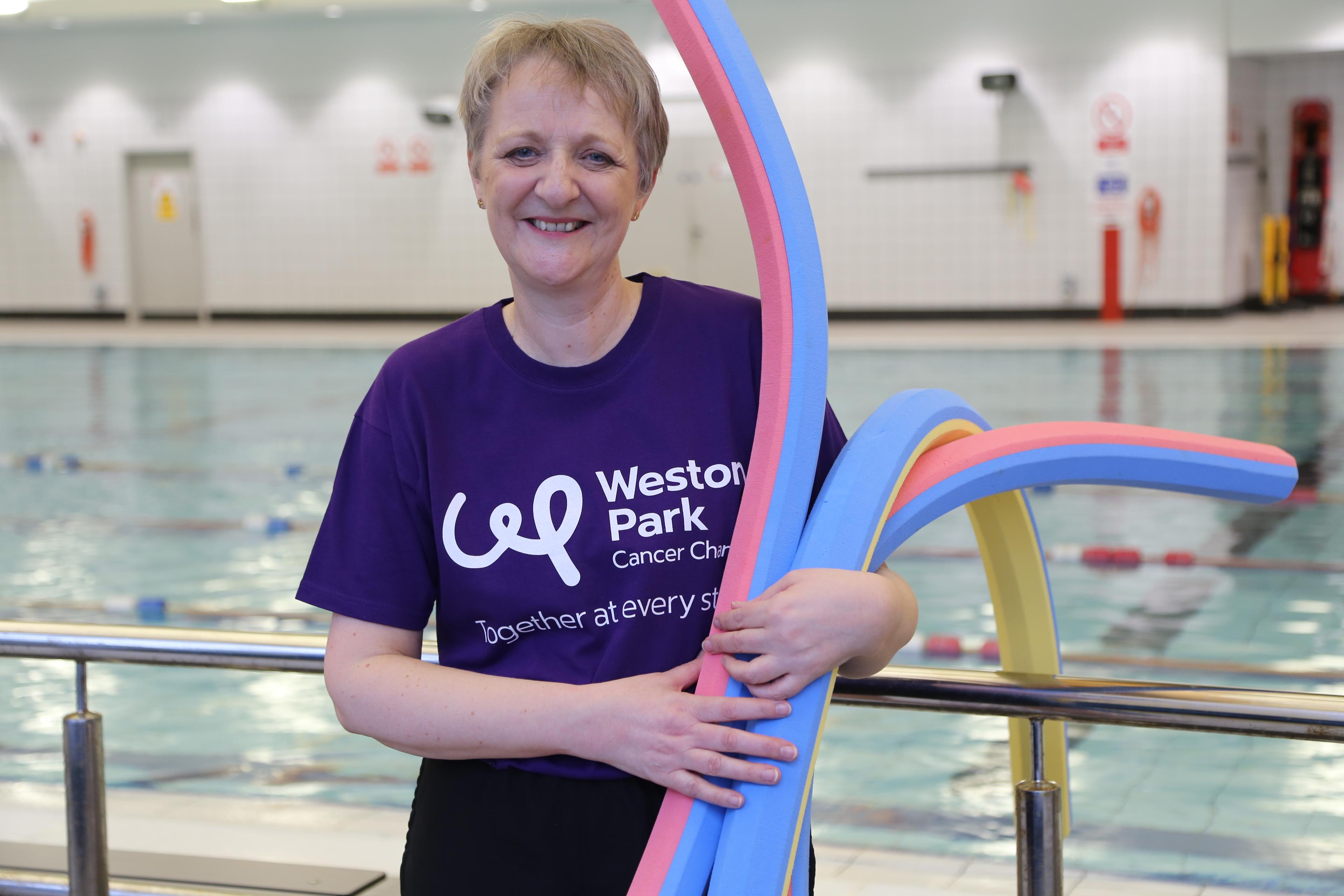 A smiling woman wearing a weston park cancer charity t shirt and holding swim aids is standing in front of a swimming pool