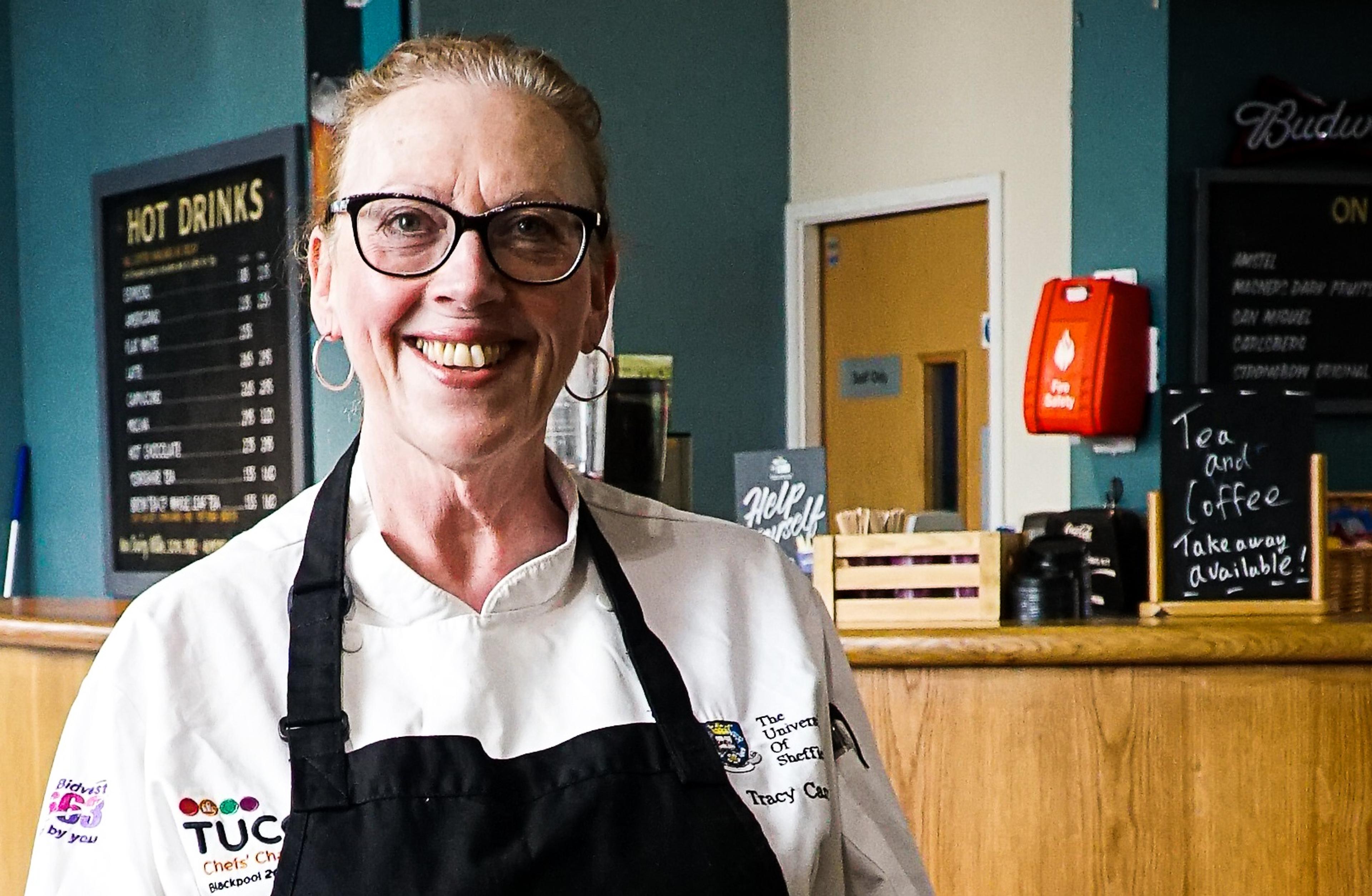 A woman stands in front of a restaurant bar, indoors, wearing an apron and facing the camera, smiling. The woman is University of Sheffield Executive Chef, Tracey Carr.
