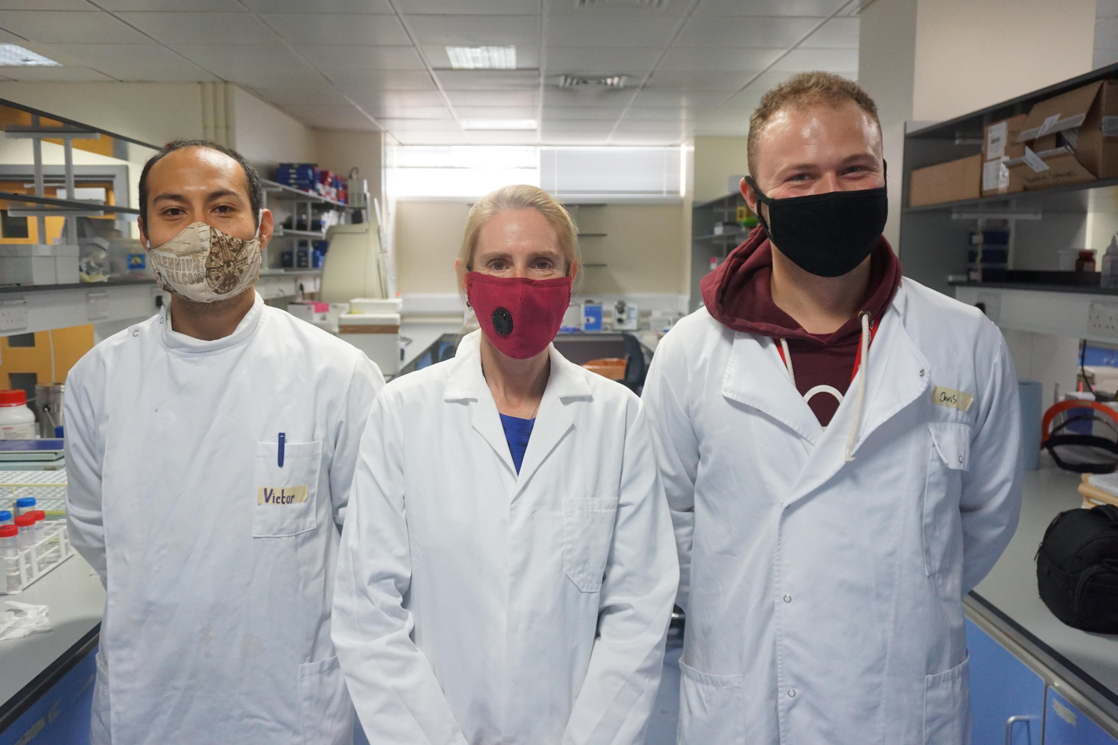Three scientists wearing face masks and white lab coats are shown standing in a laboratory