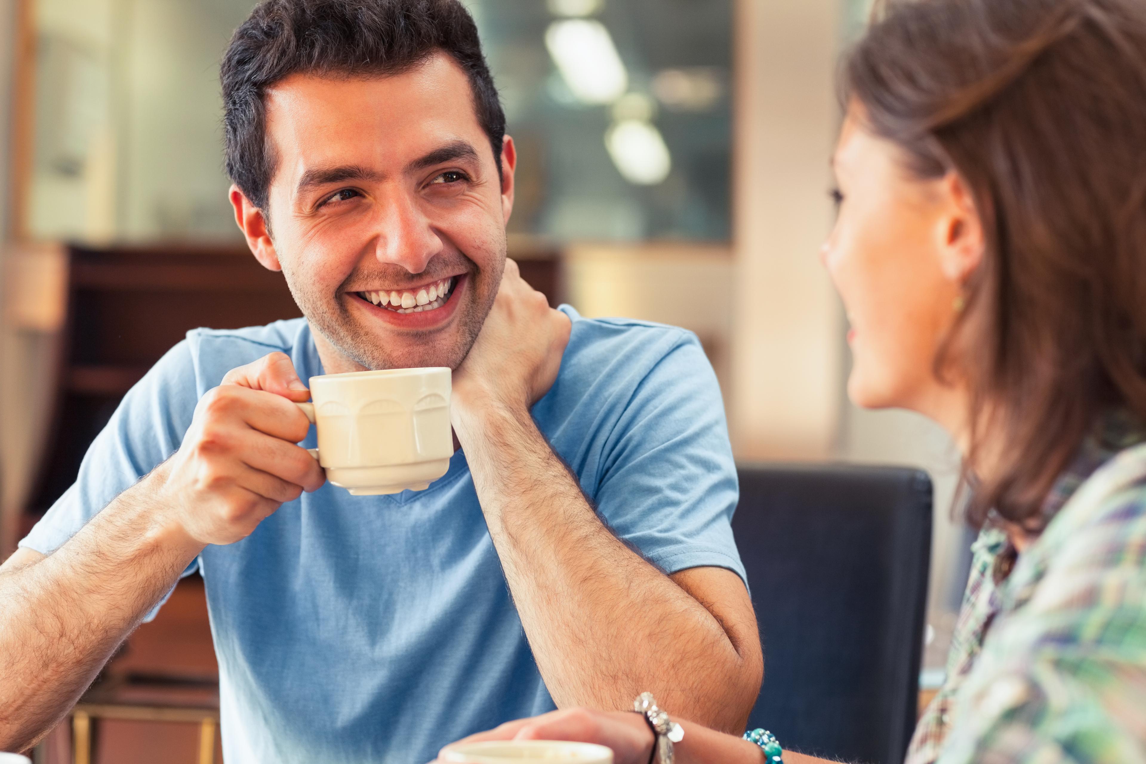 A man is holding a coffee mug up as he smiles at a woman who is talking to him