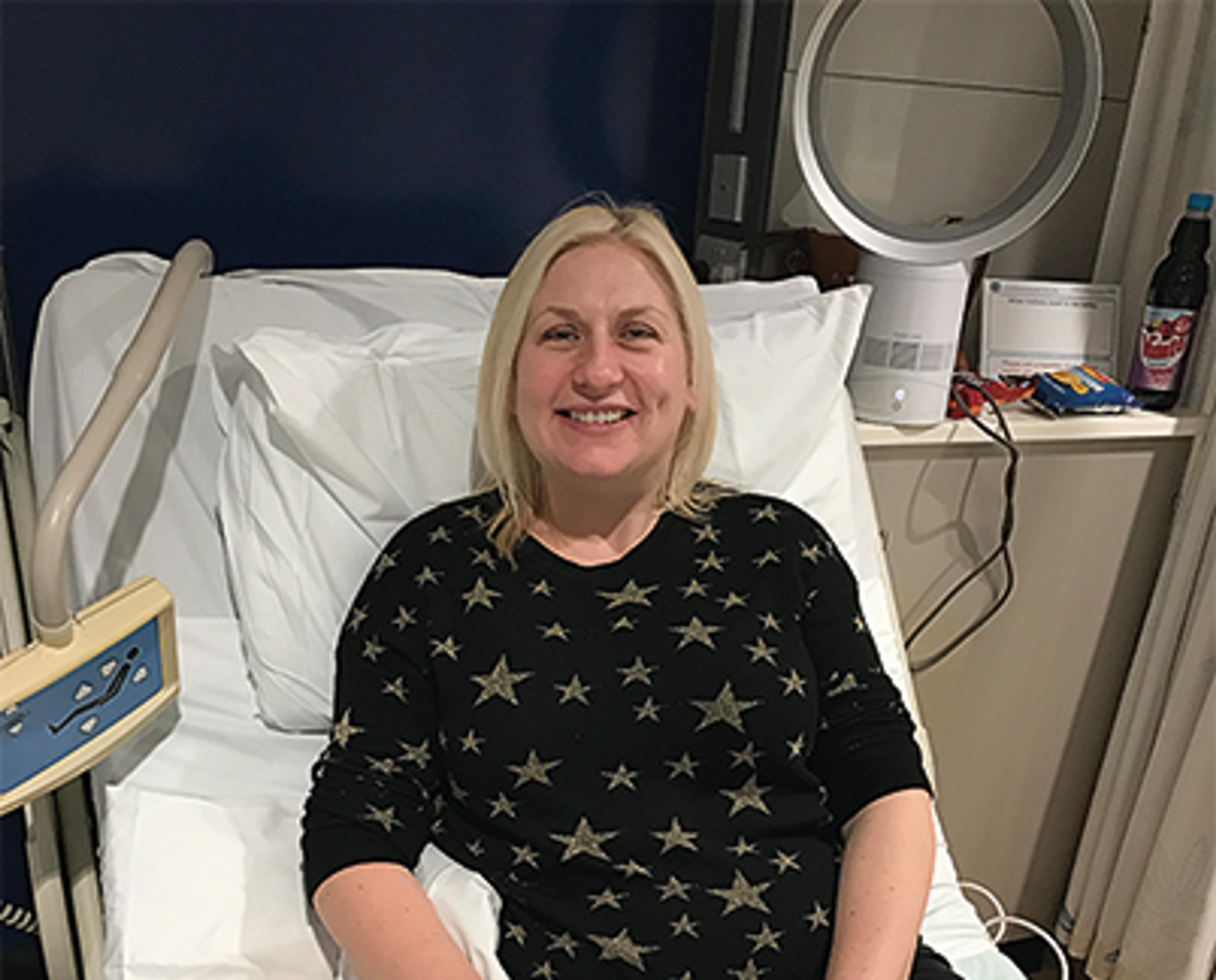 A lady, Amanda Horsman, sits in a hospital bed facing the camera, with her arm attached to a drip.