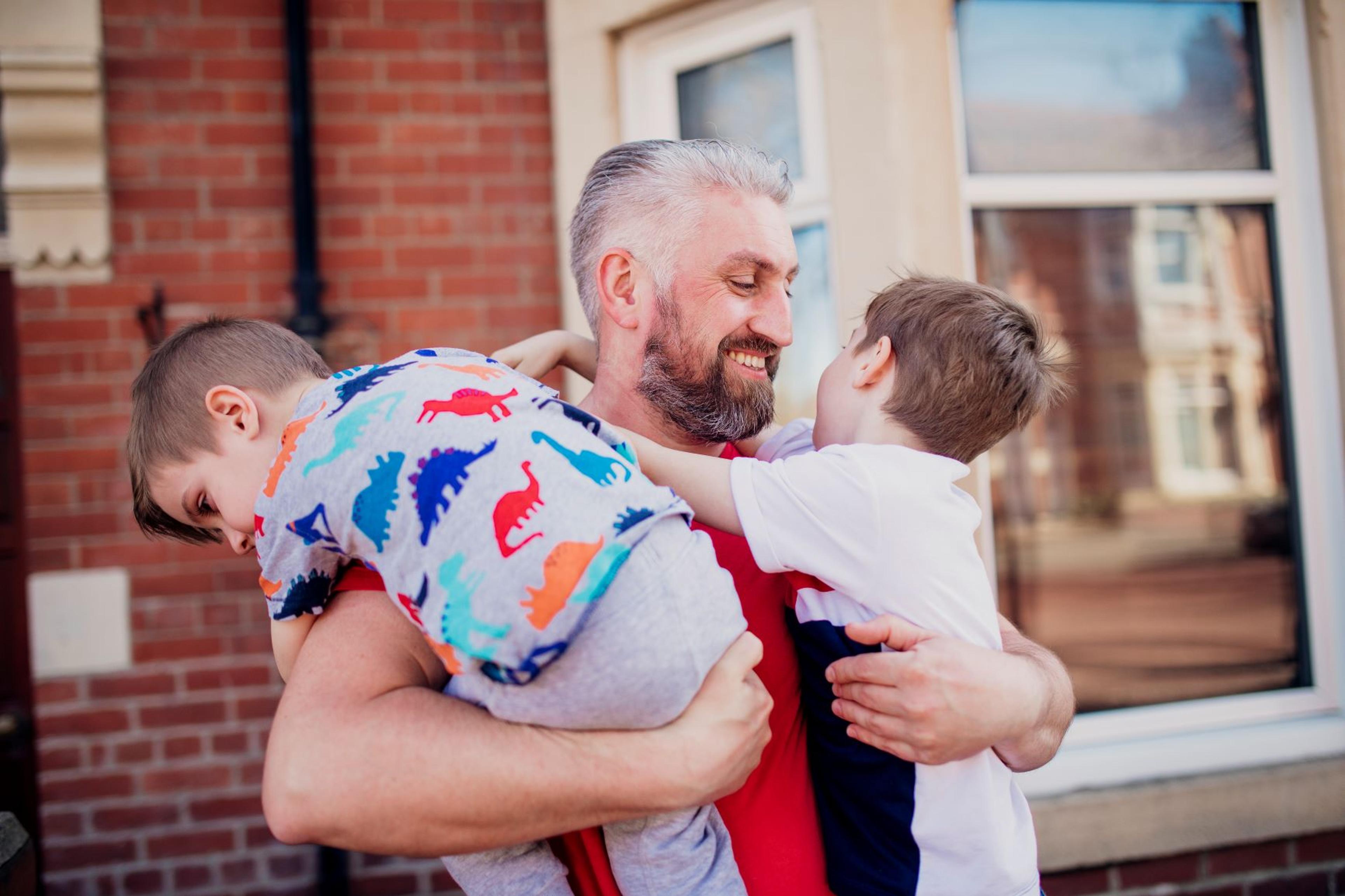 A man stands outside a house, in his arms are two young boys hugging him. He is smiling at them.