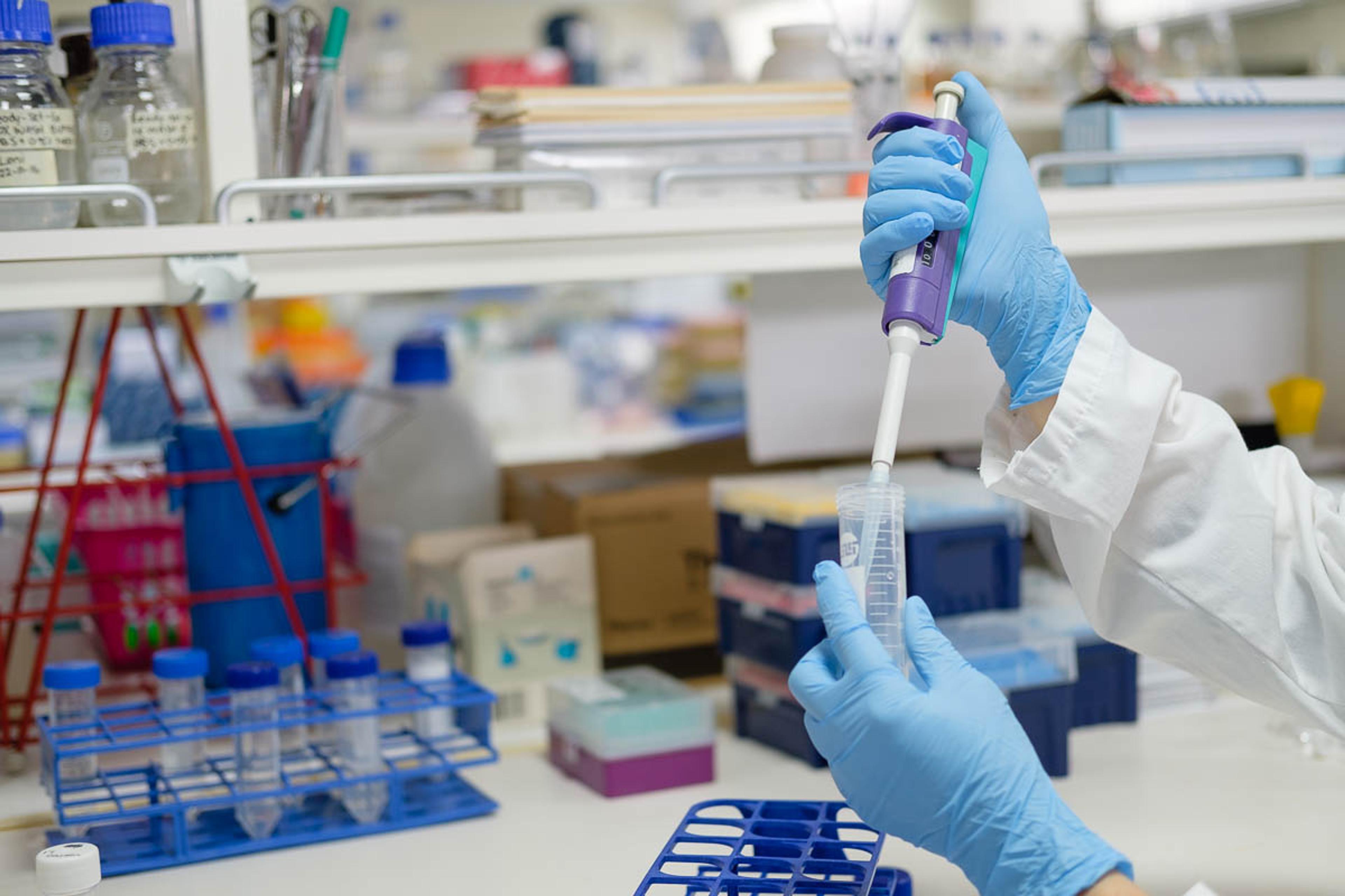 Hands with blue gloves on using a syringe to fill a test tube on a laboratory bench.