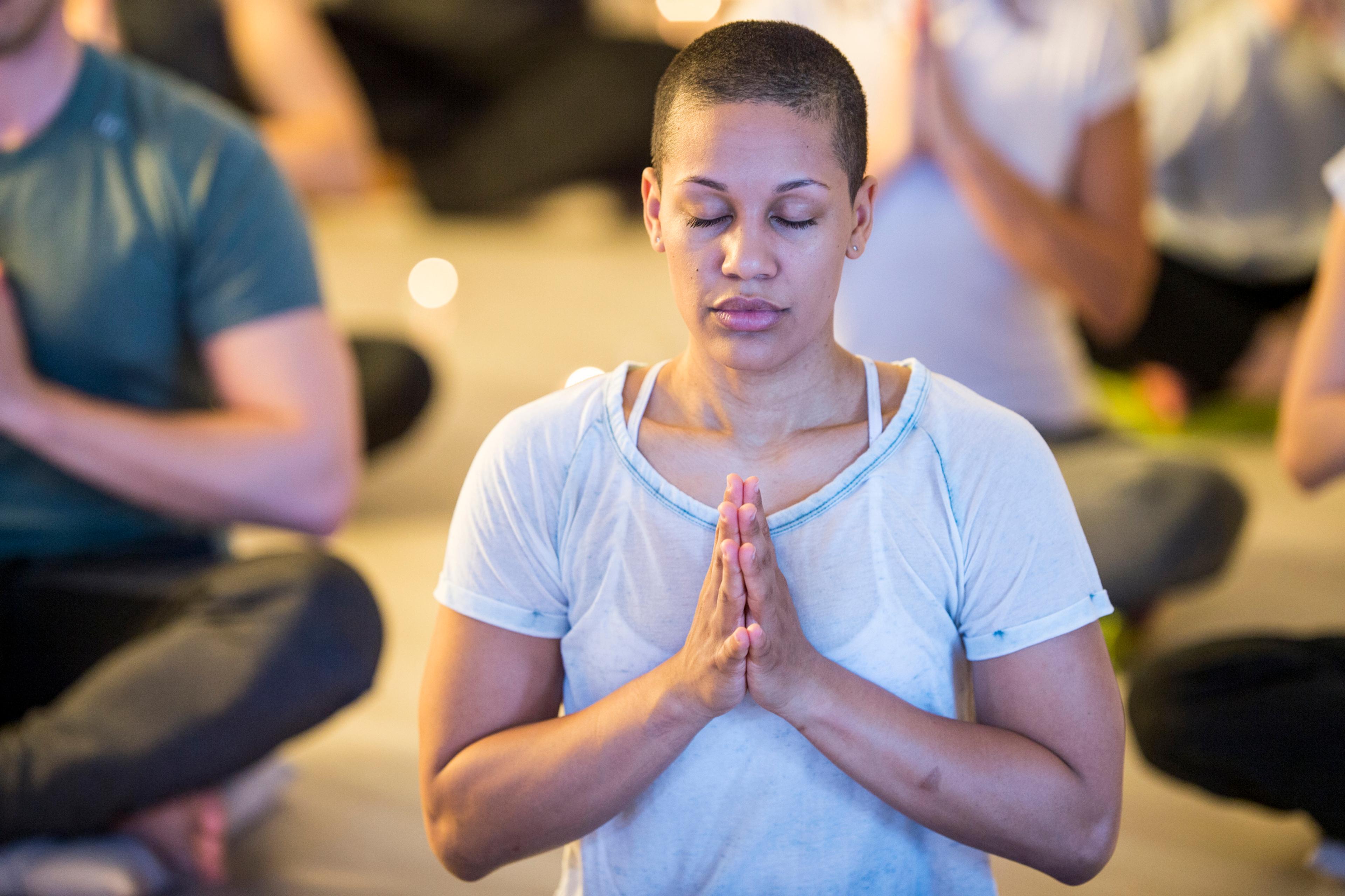 A woman with her hands in prayer position is sat cross-legged in a room of people meditating with her eyes closed.