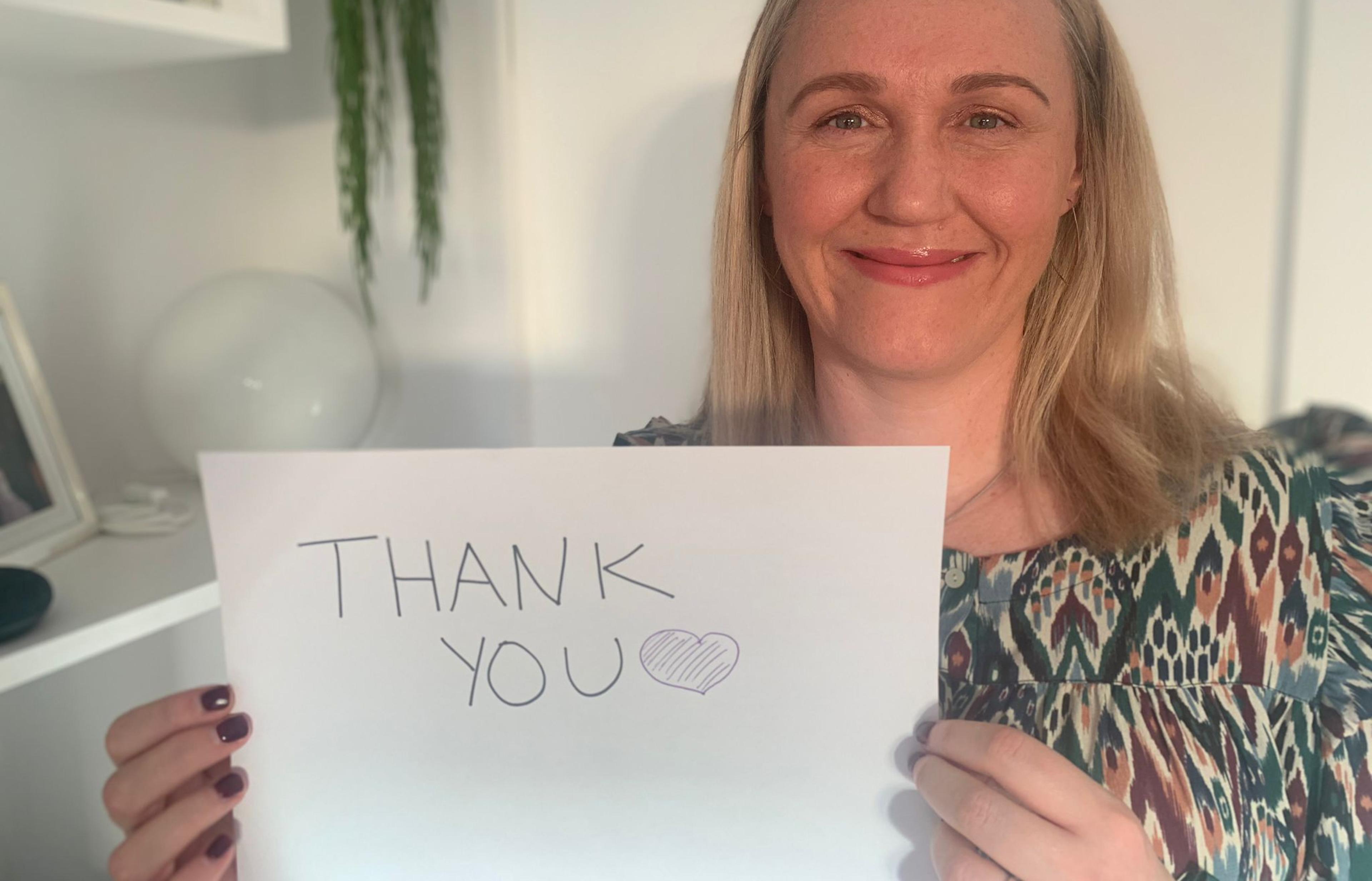 A blonde-haired lady - Weston Park Cancer Charity CEO, Emma Clarke - sits smiling while holding up a white piece of paper which reads "Thank you" with a heart drawn afterwards.