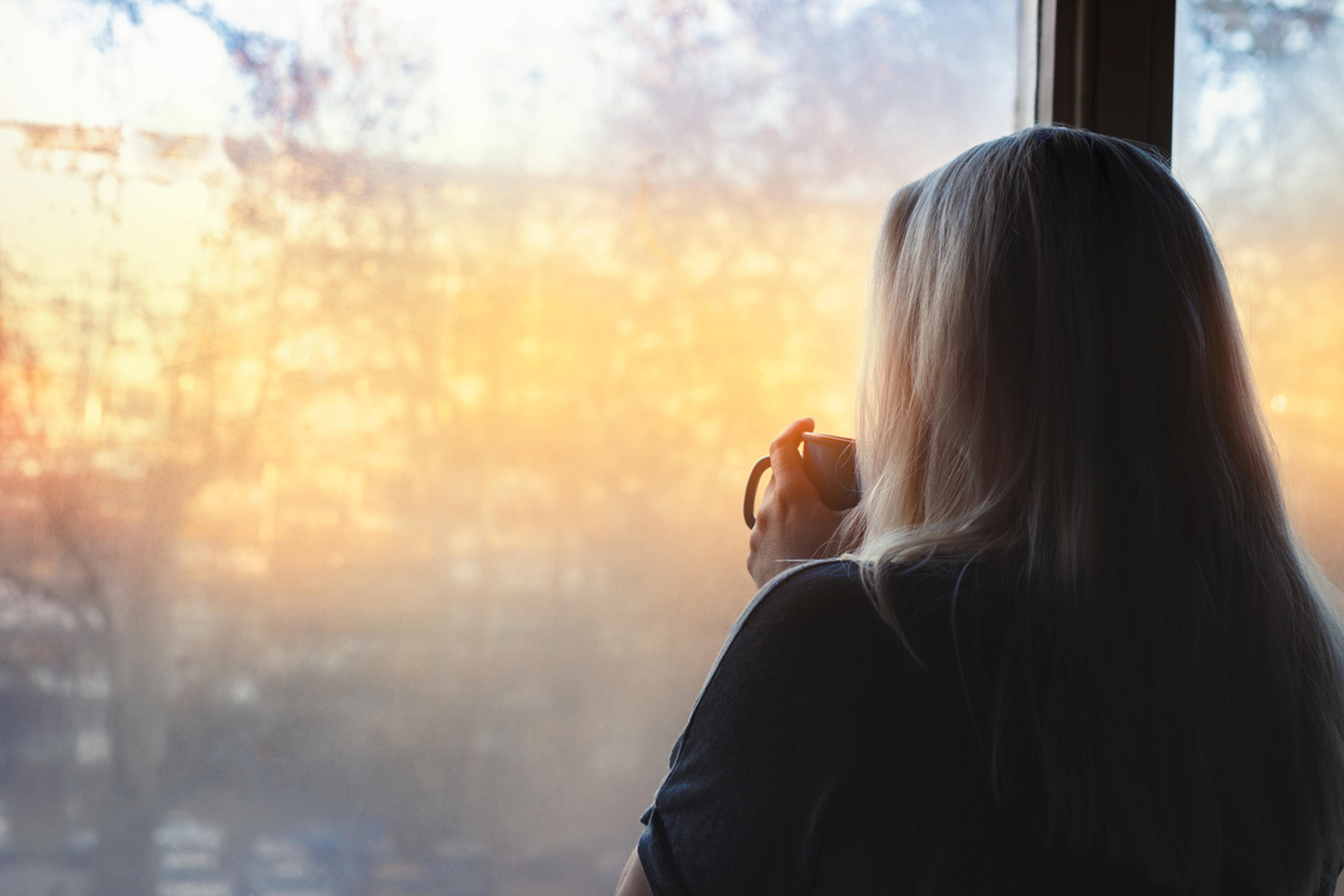 A woman looking out of a window holding a mug in her hand. Through the window is a hazy scene of winter sunshine.