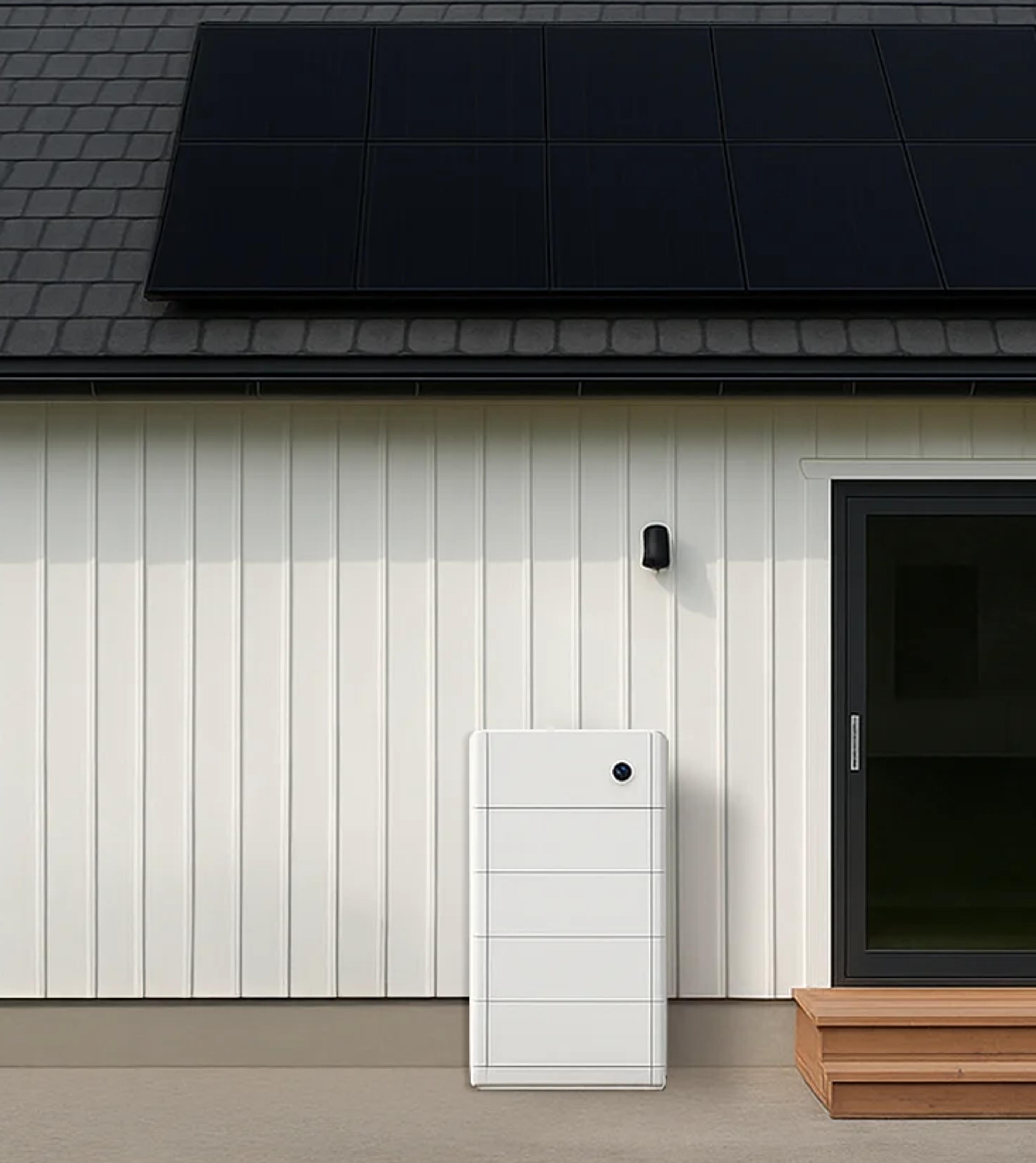 Et modulært batteri, montert på ytterveggen av et hvitt hus med solcellenpaneler på taket