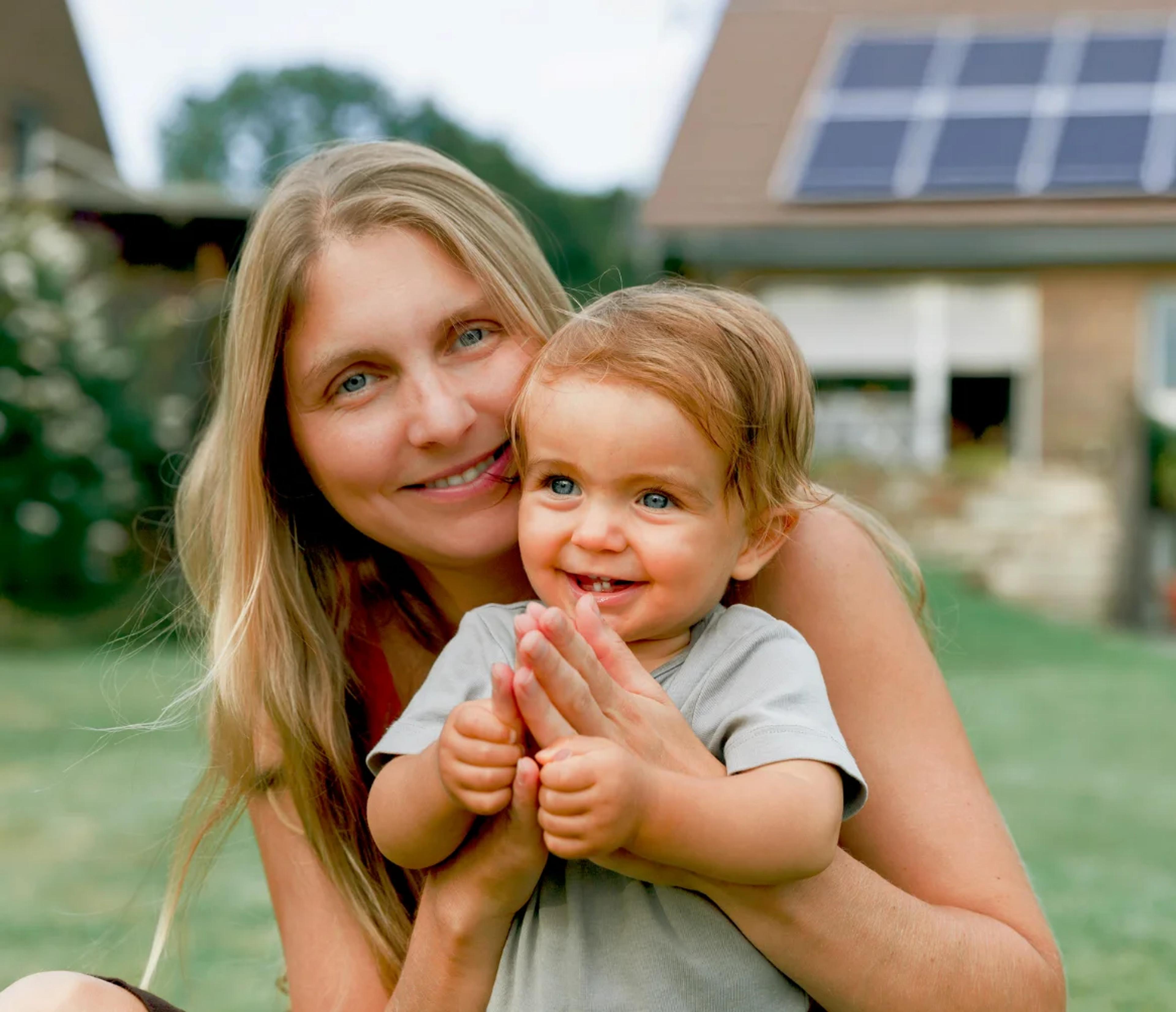 Mutter mit einem Kind im Vordergrund und ein Haus mit PV im Hintergrund