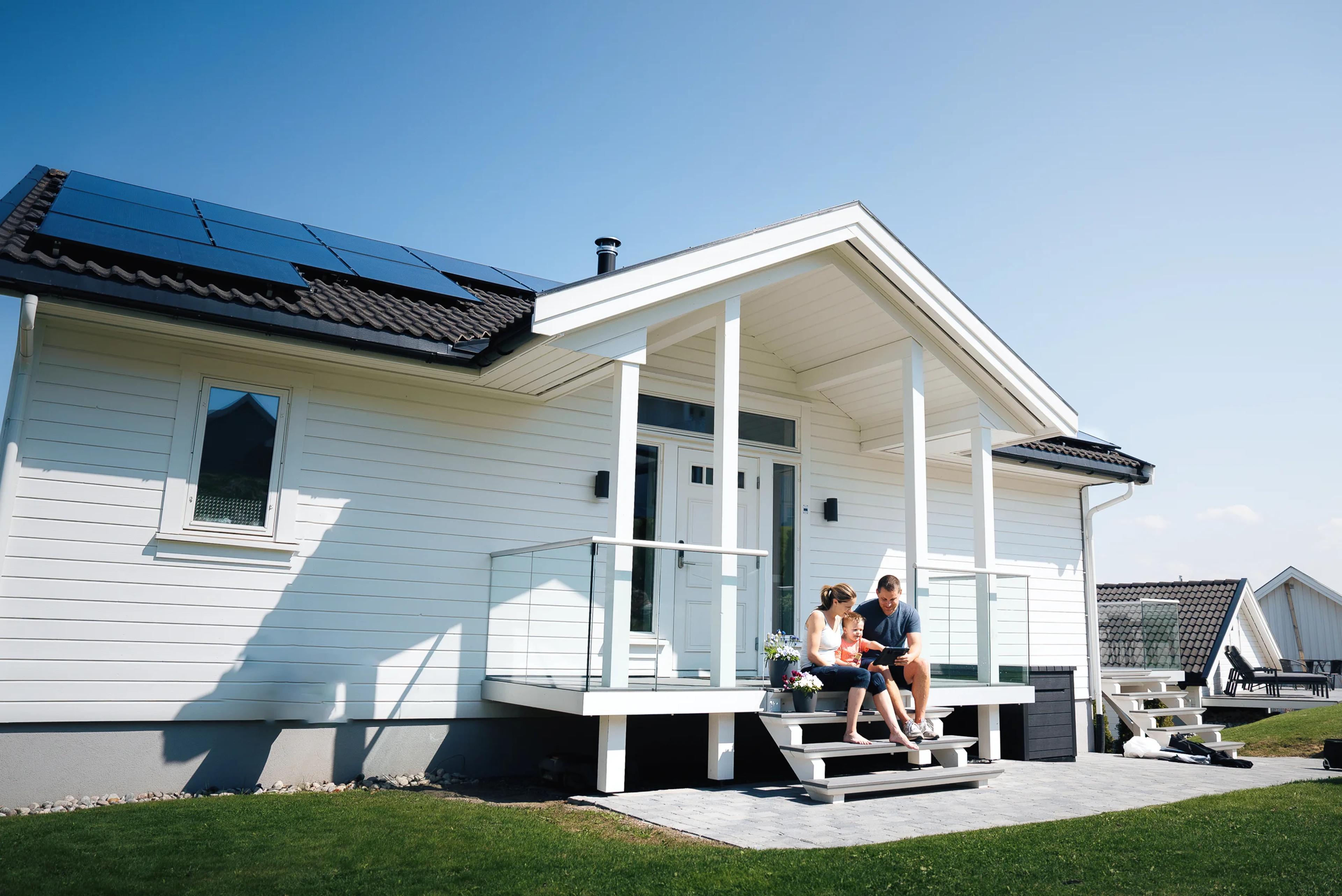 Et foreldrepar med et lite barn sitter på trappen til et hus som har solcellepaneler på taket