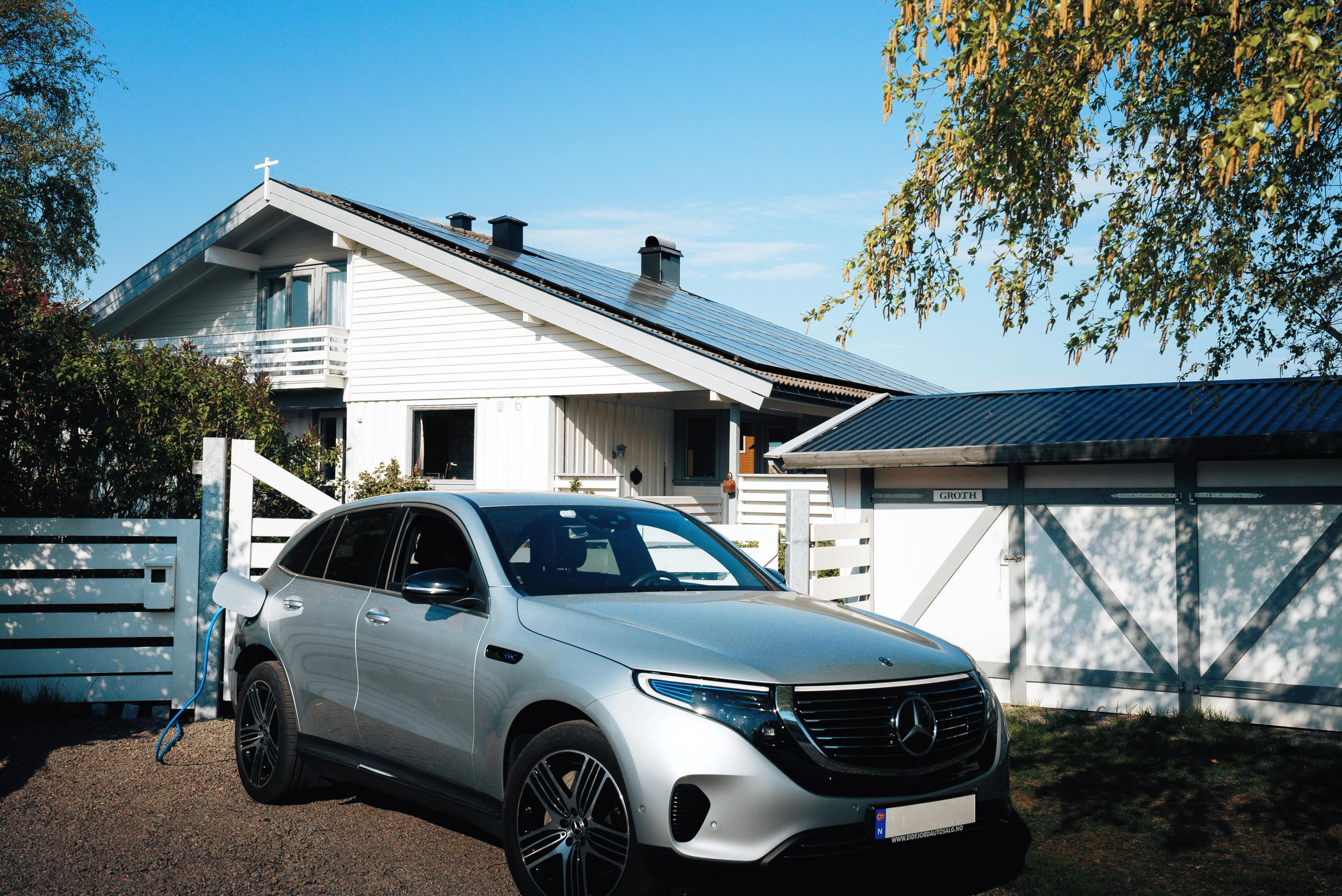 En sølvgrå bil lader utenfor et hvitt hus med solceller på taket
