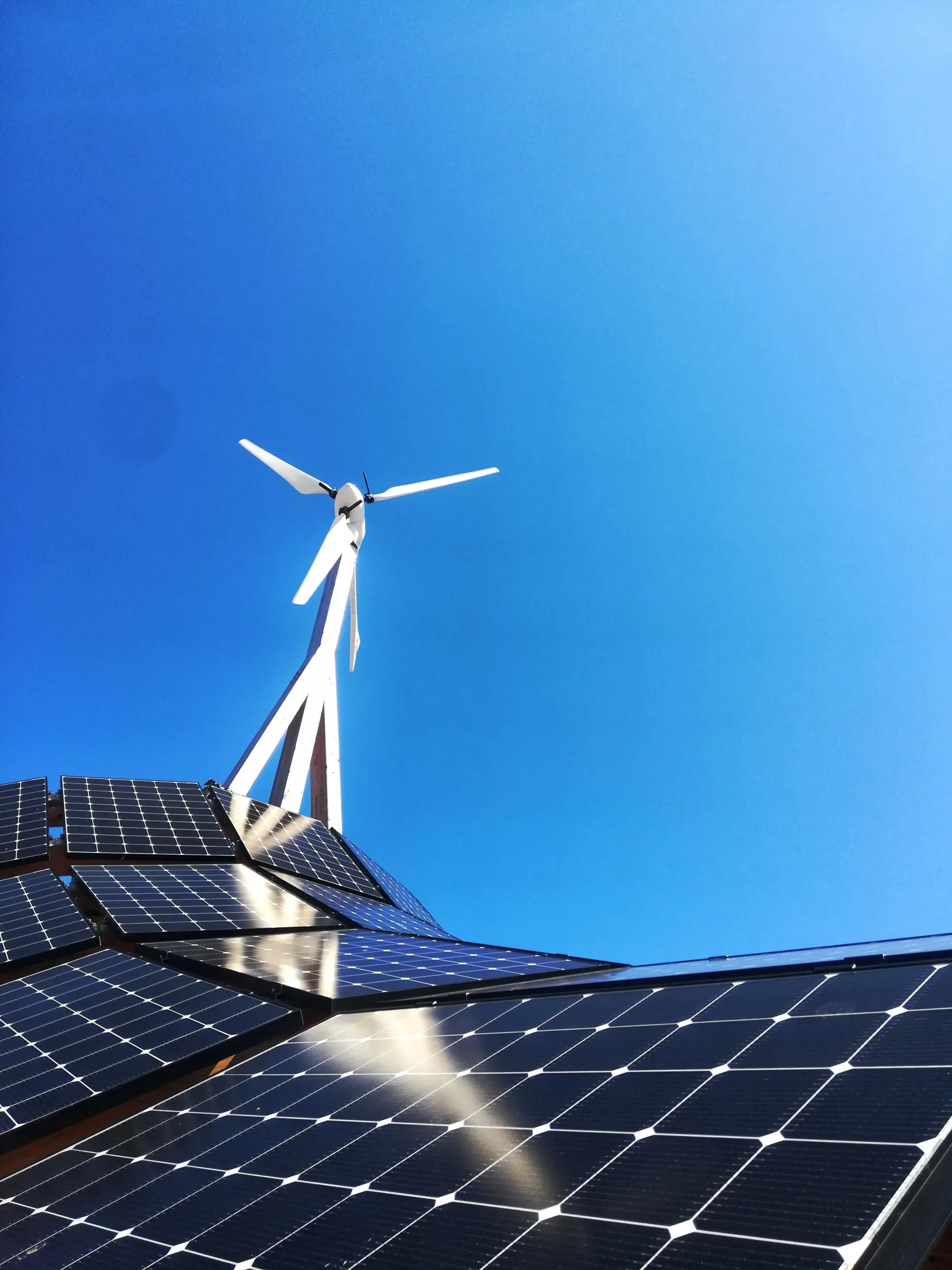 Solar panels on a roof, leading to a wind turbine. The panels are set against a clear blue sky.