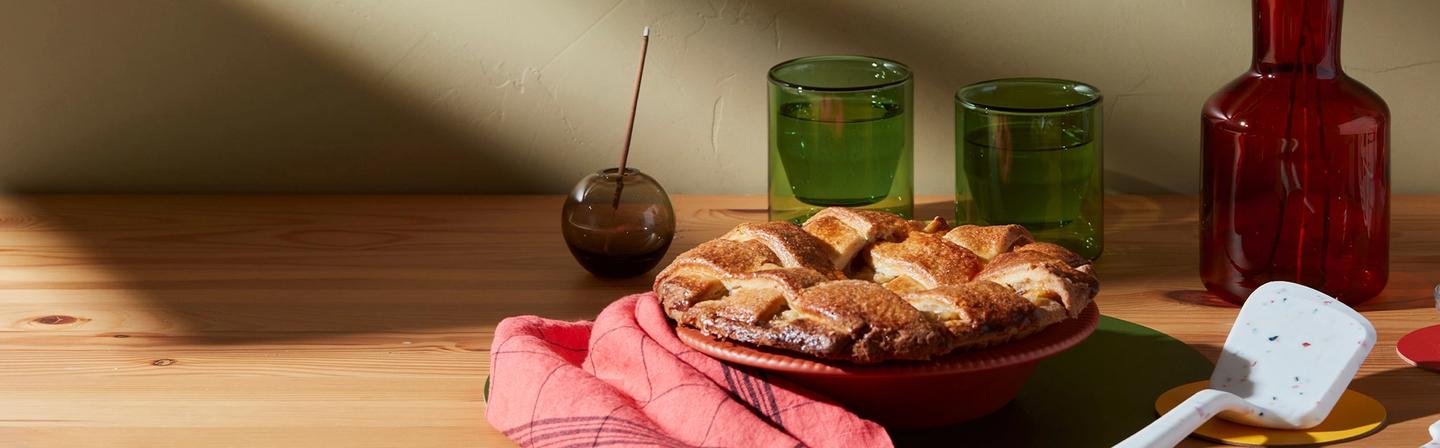 A golden apple pie sitting on a wooden counter with a GIR spatula next to it with glasses in the background.