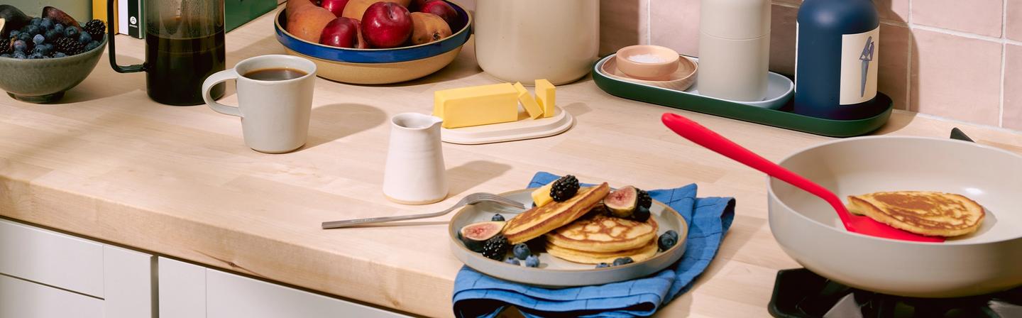 A counter view of fruit bowls, plates with pancakes apancakes and nd GIR kitchen utensils.