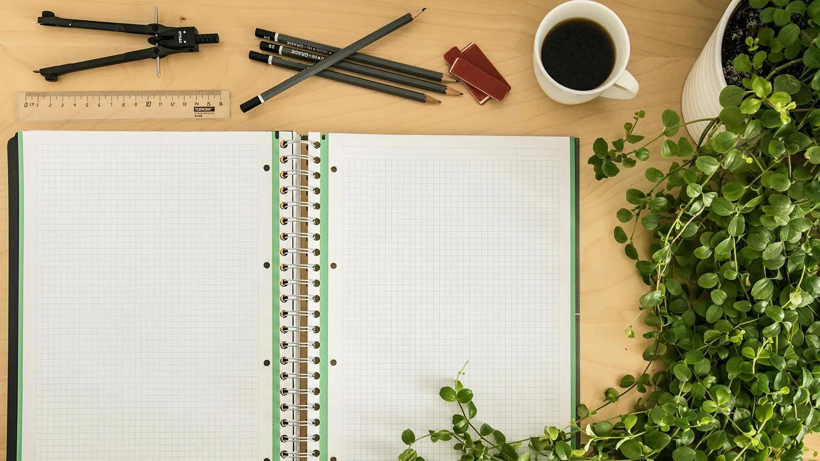 Overhead view of a study desk with an open blank grid notebook, coffee cup, plant, pencils, compass, and ruler, ready for studying or note-taking.