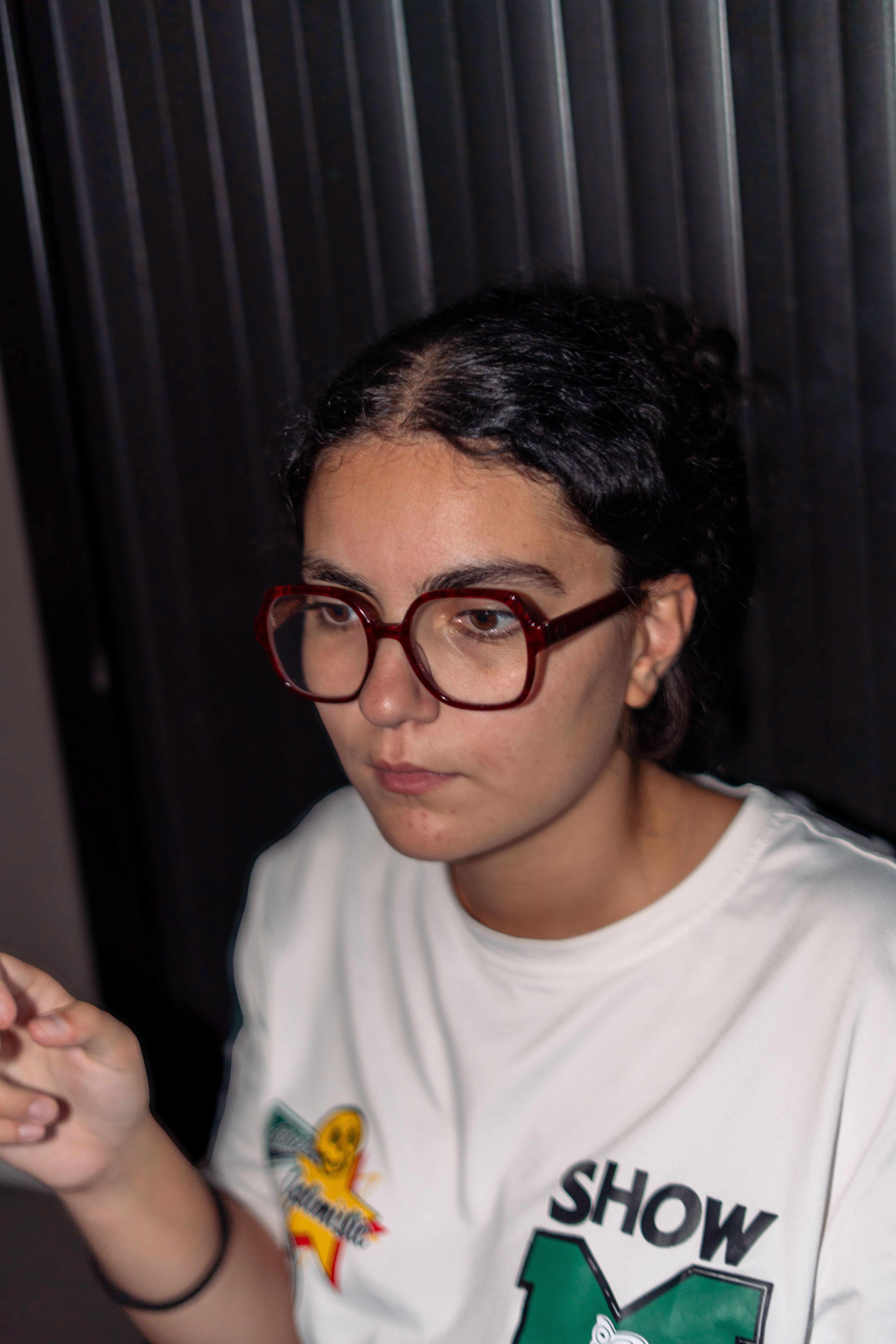 Portrait d'une jeune femme à lunettes rouges, détournant le regard, sur un fond de rideau sombre