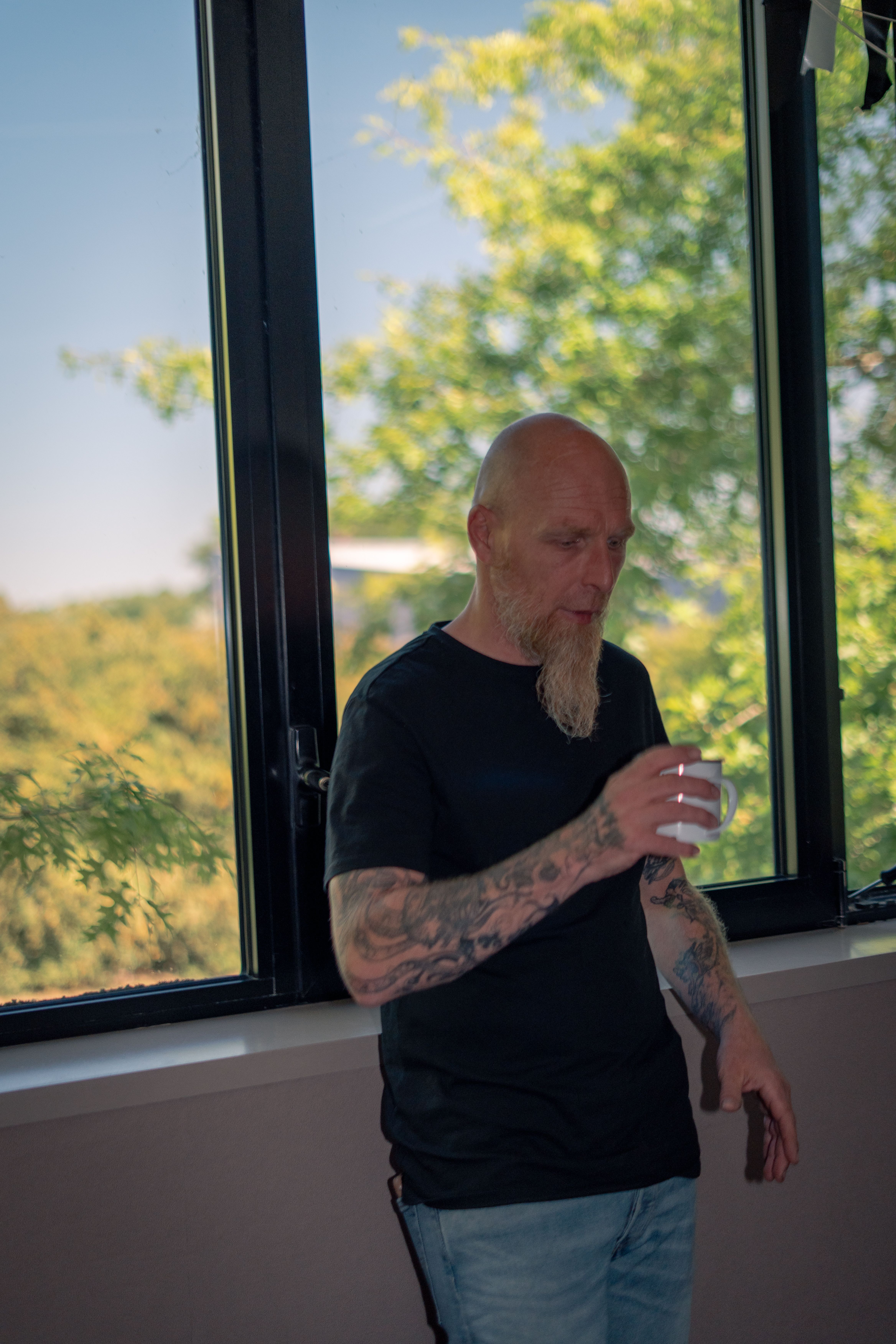 Un homme tatoué tient un gobelet devant une fenêtre ouverte sur les arbres, entre deux prises