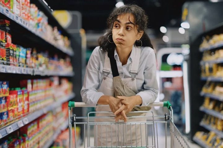 A woman leans on a shopping cart in a grocery store aisle, looking worried