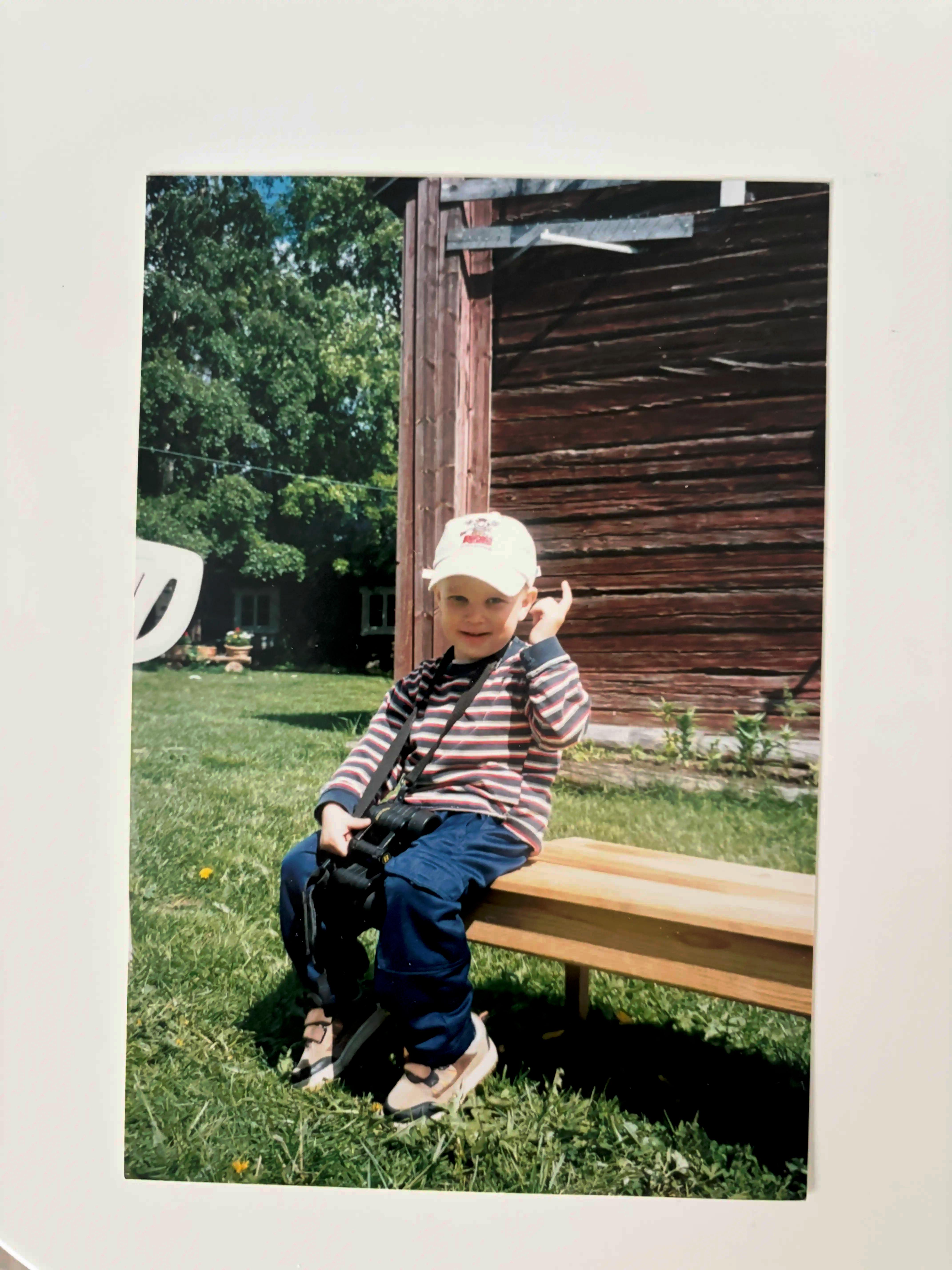 A photograph showing Aake Juvani as a child sitting on a bench, wearing a white cap