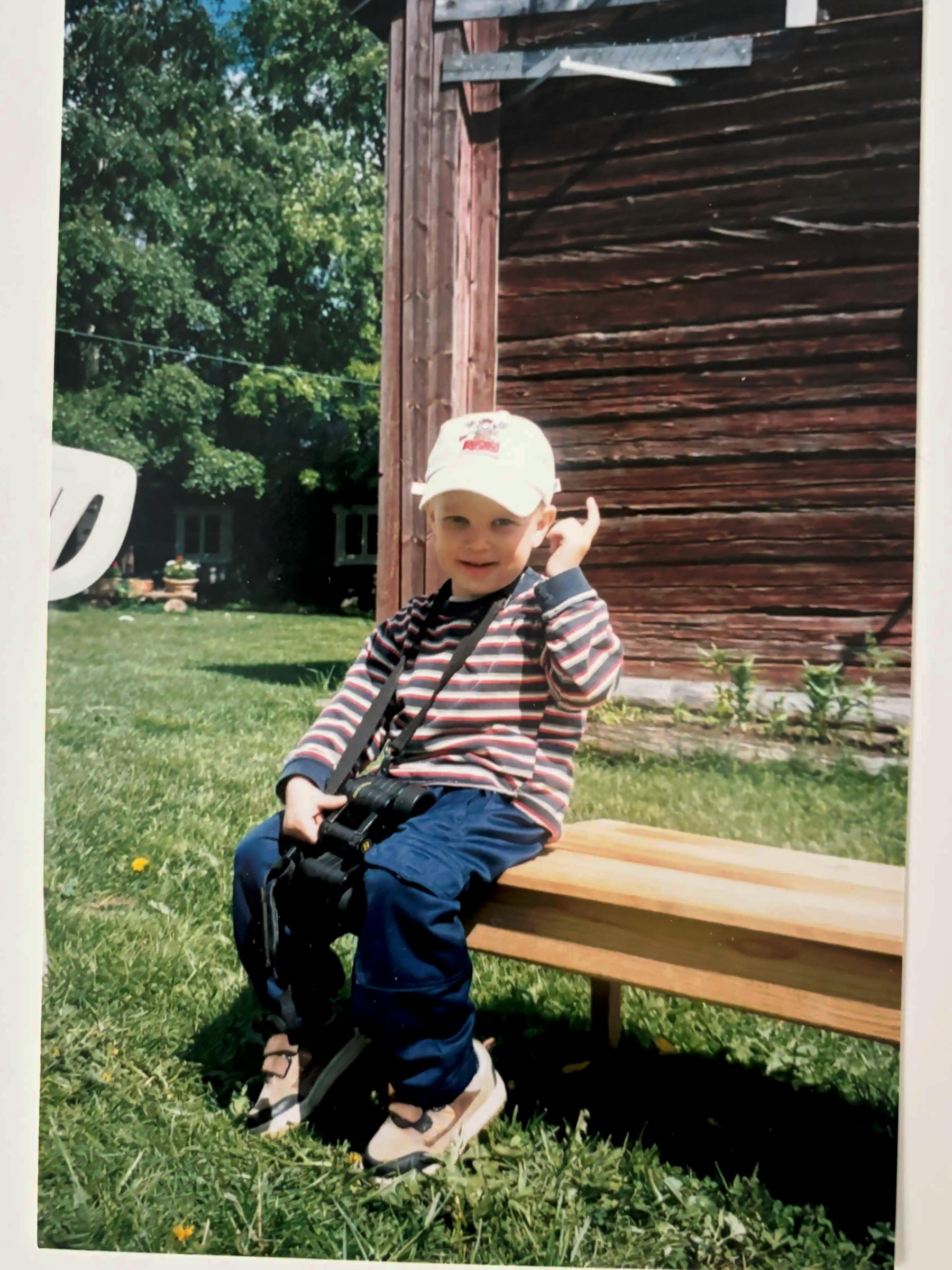 A photograph showing Aake Juvani as a child sitting on a bench, wearing a white cap