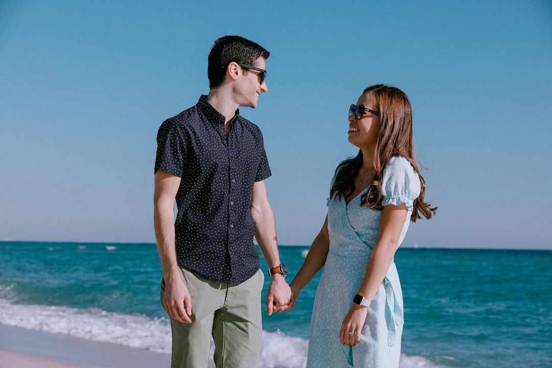 Mark Rini stands on the beach in front of the ocean, holding his fiancée's hand. The two are smiling and looking at each other.