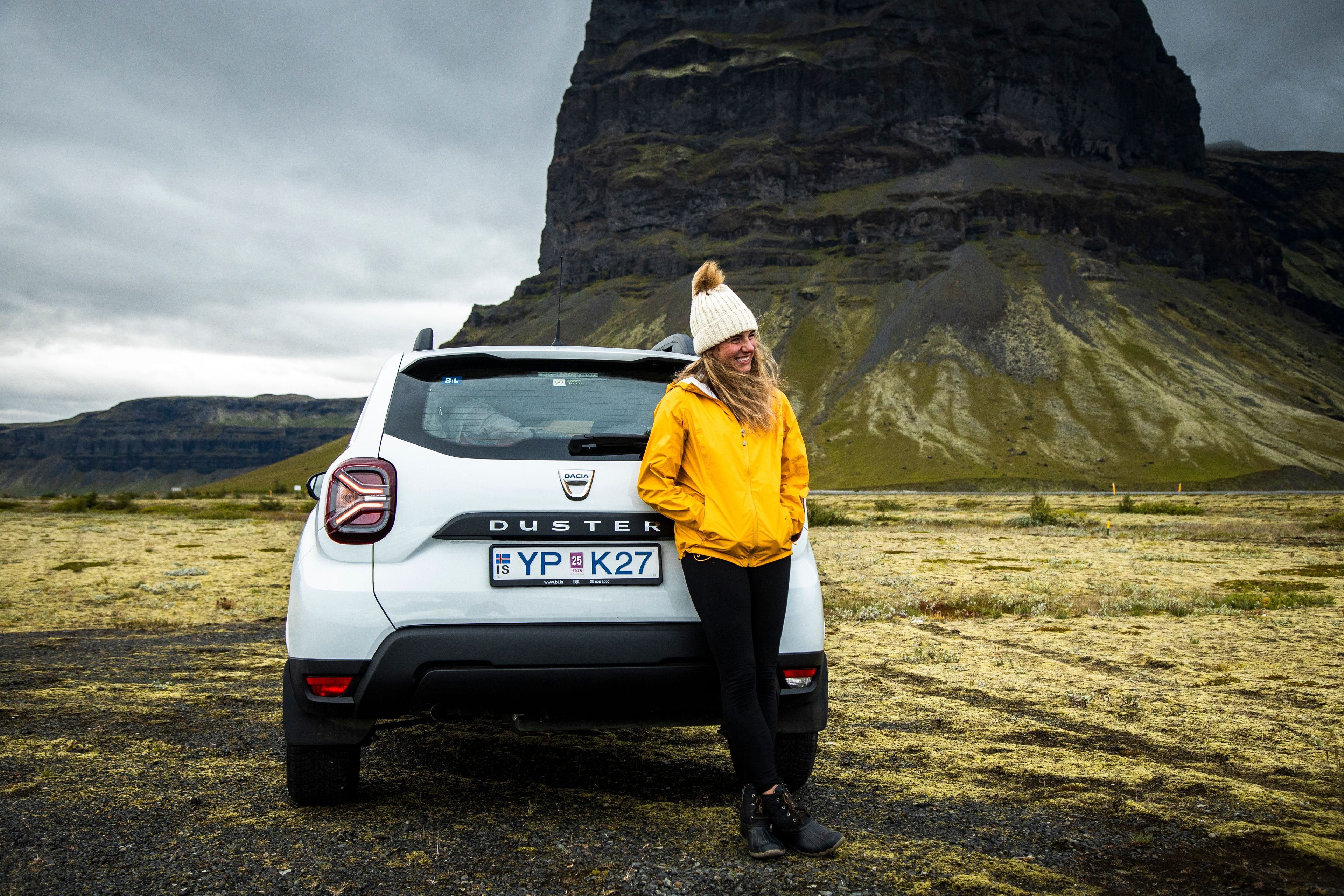 Woman wearing a yellow jacket with a smile on her face while standing next to a rental car in iceland