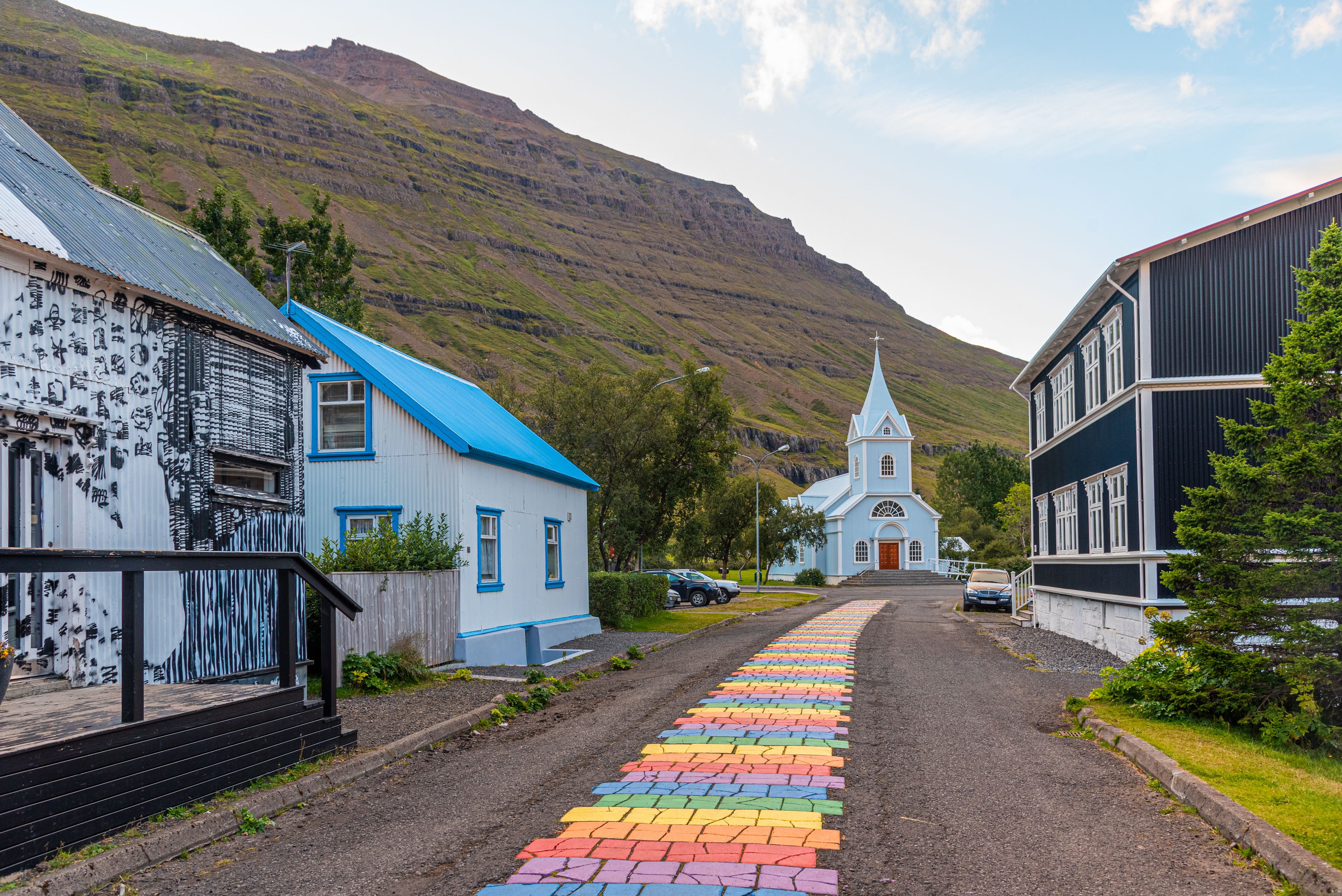 The vibrant Rainbow Road in Seydisfjordur, Iceland, stretching towards a quaint town set against mountainous scenery.