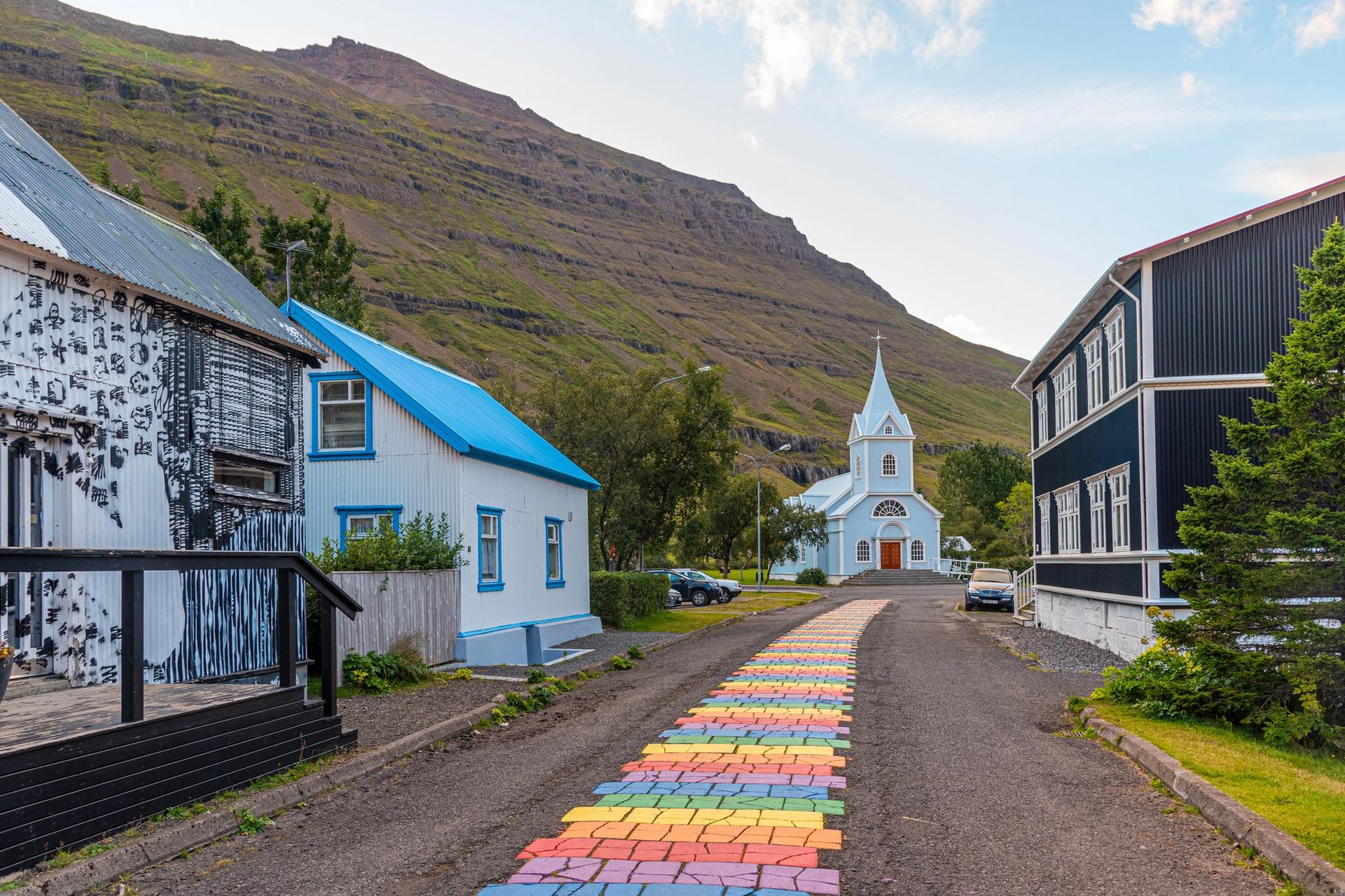 The vibrant Rainbow Road in Seydisfjordur, Iceland, stretching towards a quaint town set against mountainous scenery.