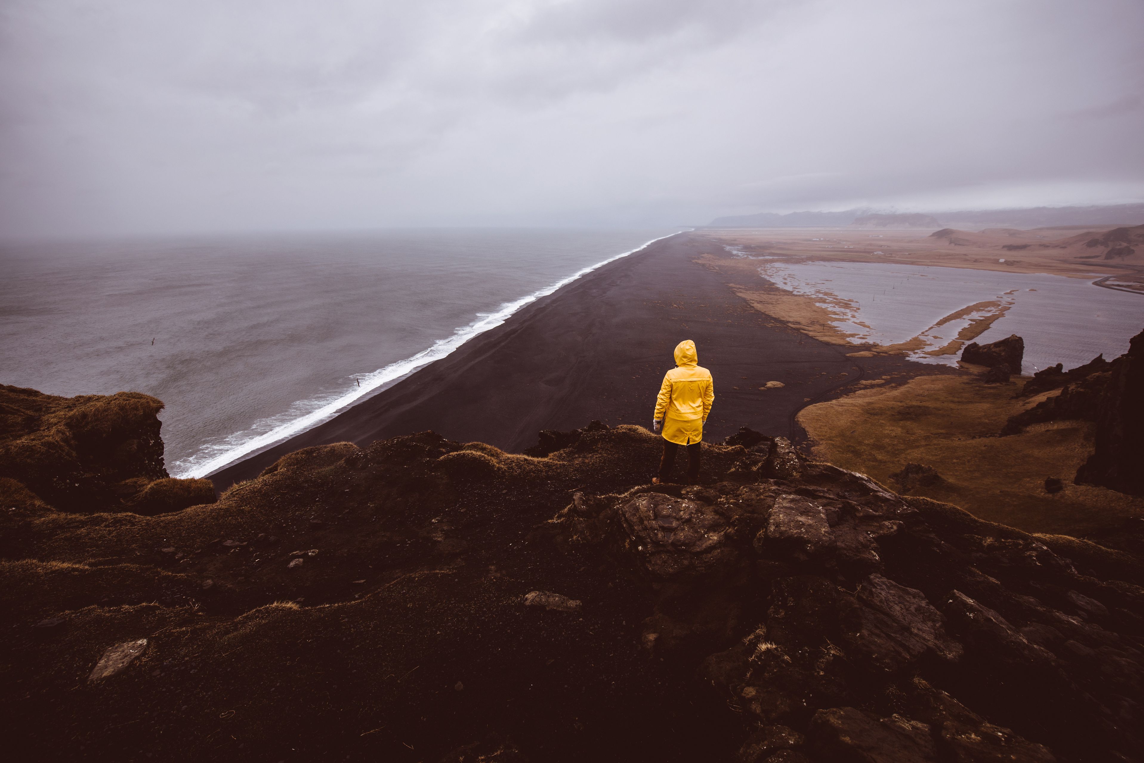 Man with a yellow raincoat on top of a cliff in front a black sand beach