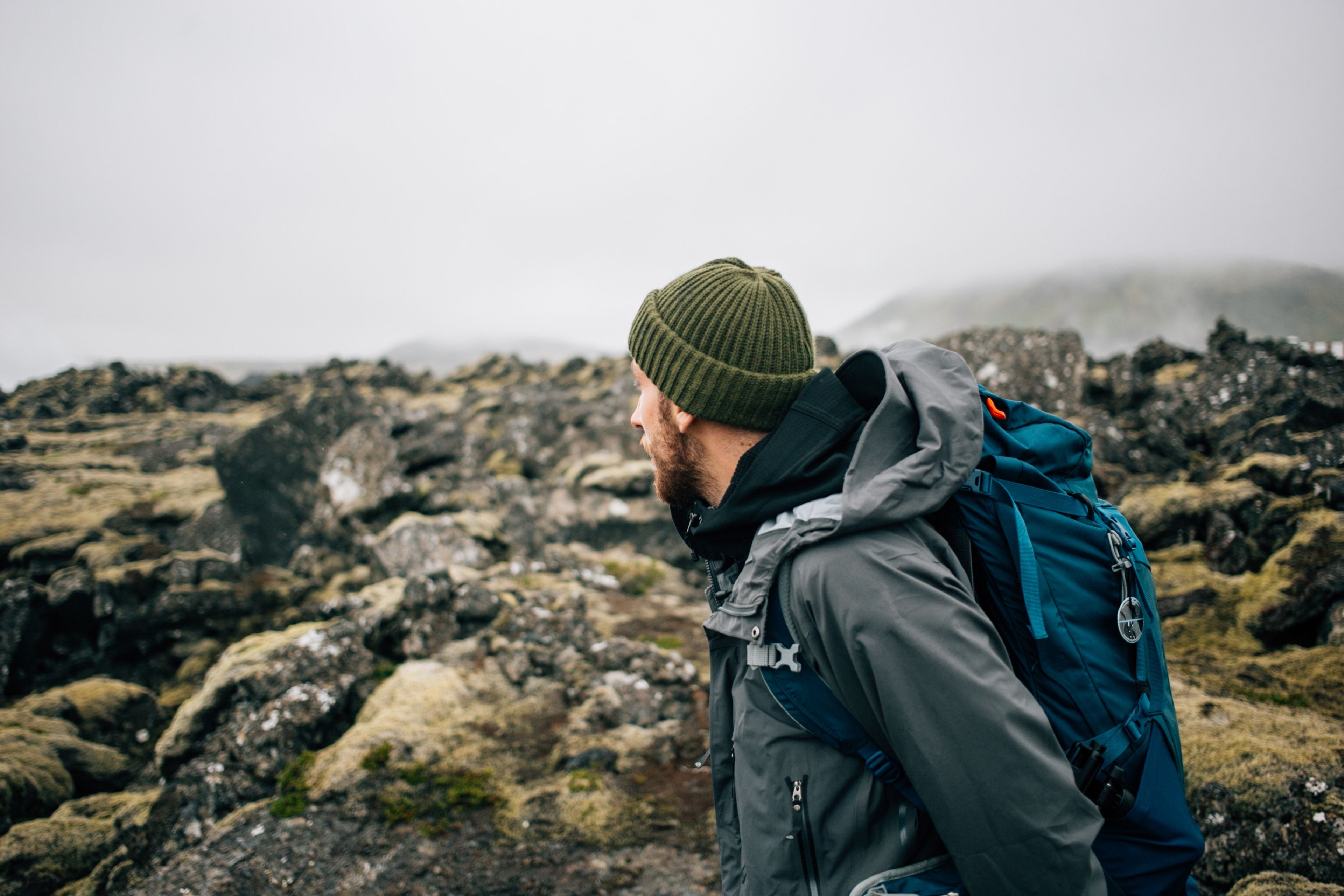 hiking trekking gear, waterproof jacket, green knit beanie and hike backpack walk through moss covered rough iceland terrain.