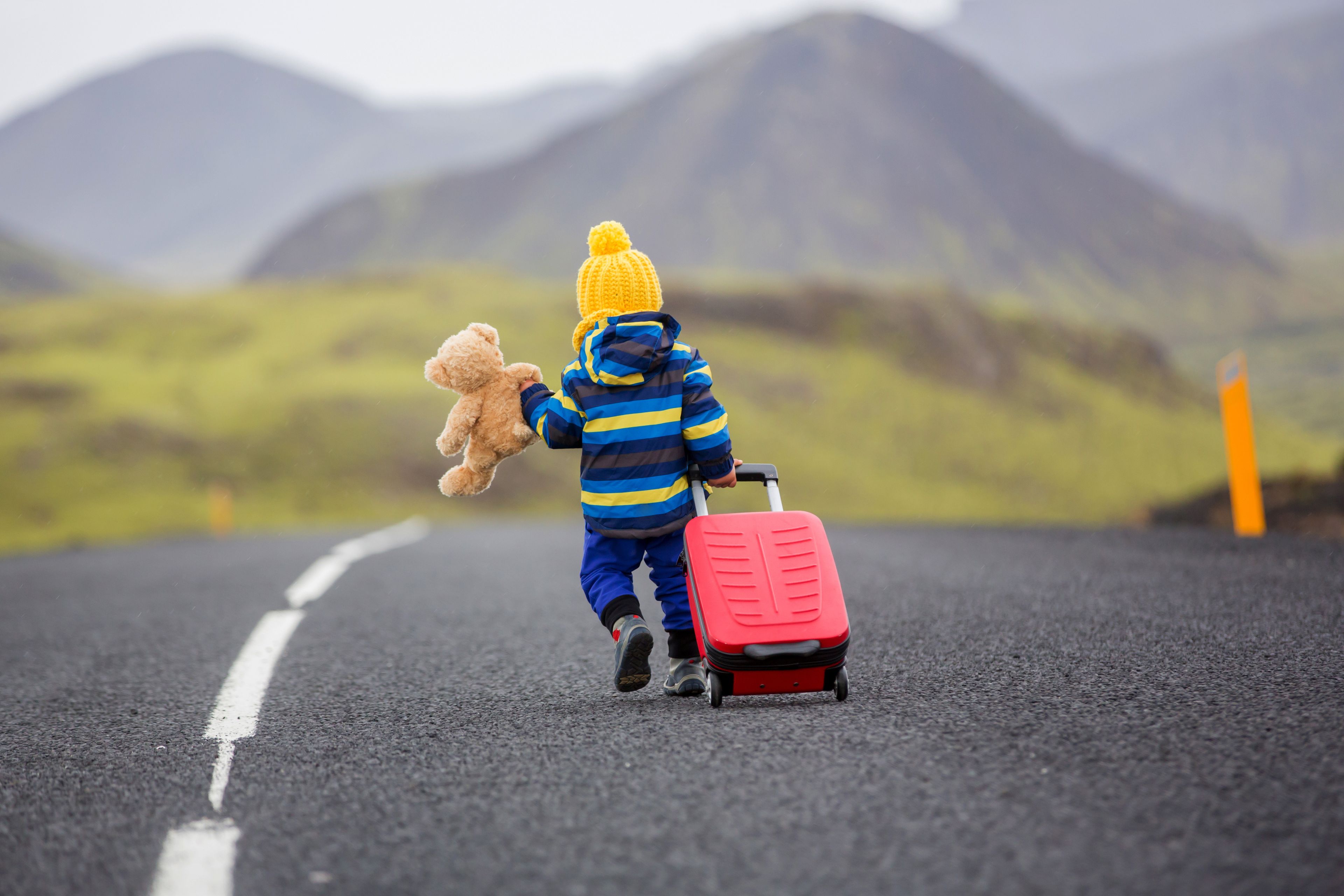 Niña pequeña con un oso de peluche una mano y una maleta de ruedas en la otra corriendo por una carretera en Islandia