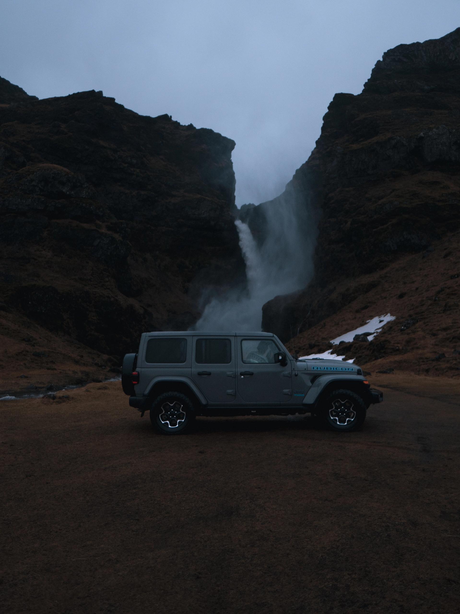 A parked dark gray Jeep Wrangler Rubicon rental car near a stunning waterfall in iceland