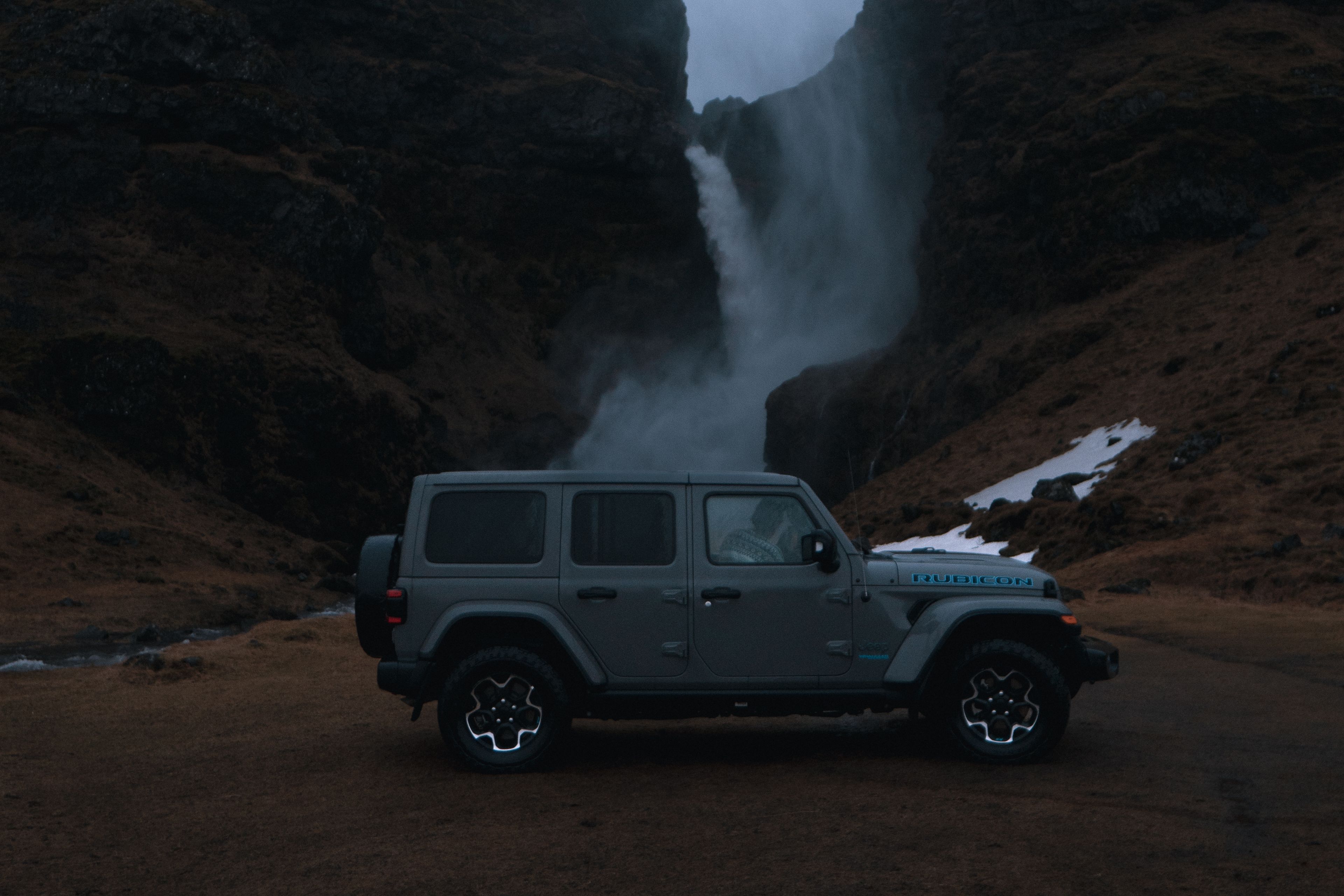 A parked dark gray Jeep Wrangler Rubicon rental car near a stunning waterfall in iceland