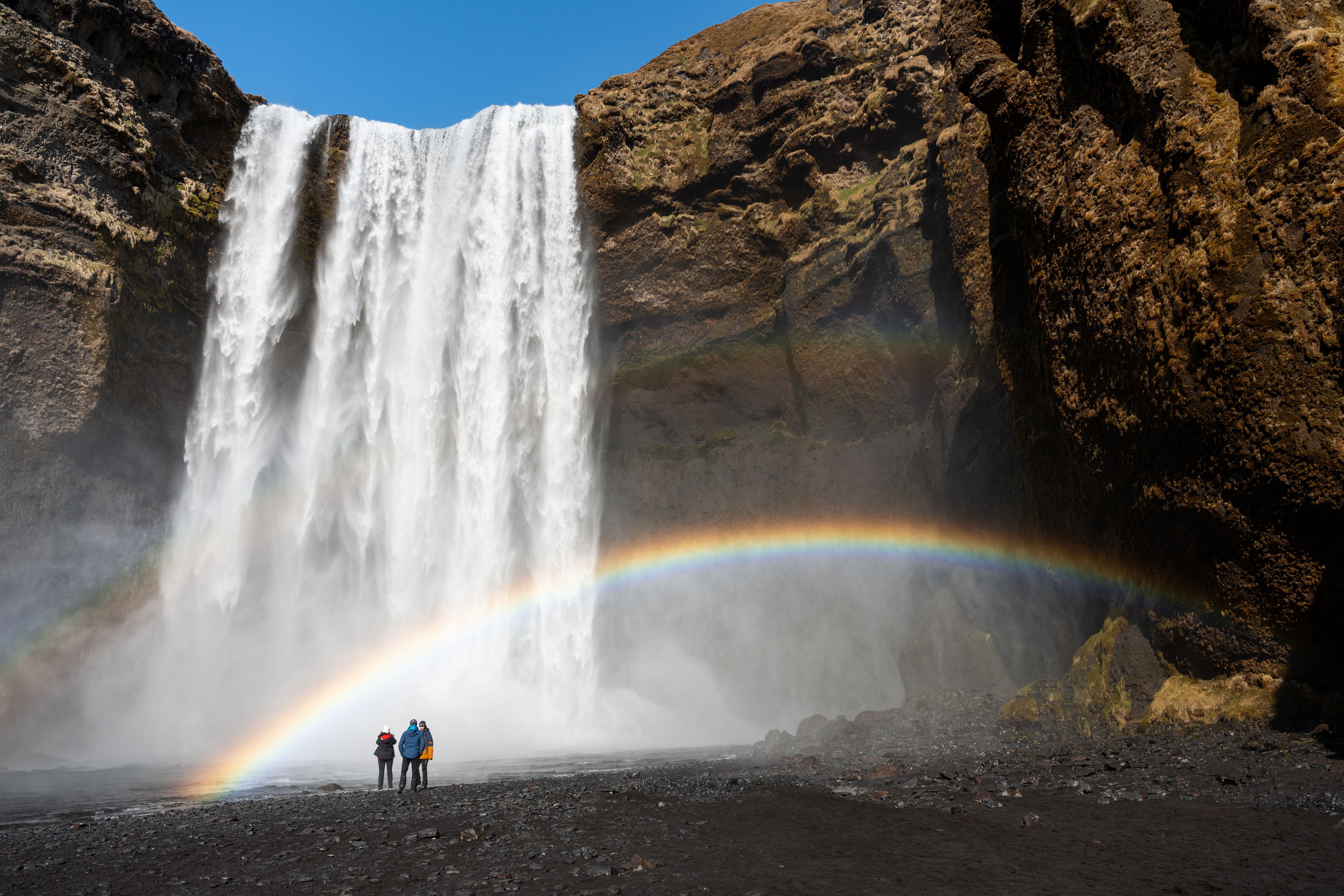 Three people in front of Skógafoss with a small rainbow 