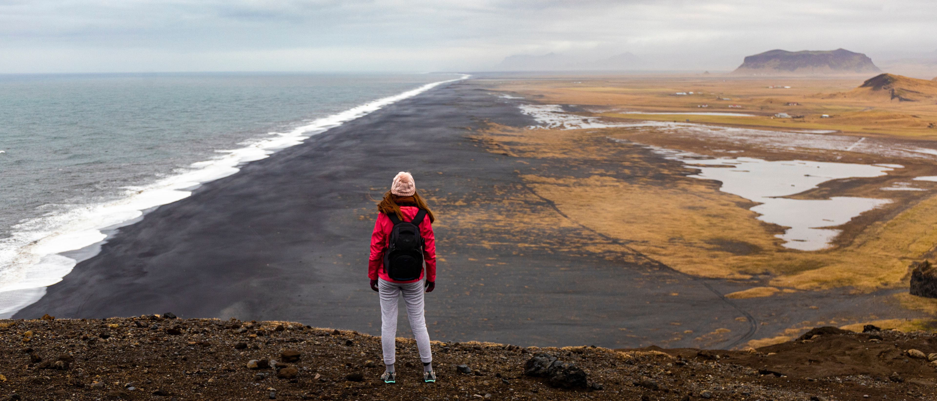 Girl in red jacket and backpack looks at a vast empty Dyrhólaey black sand beach