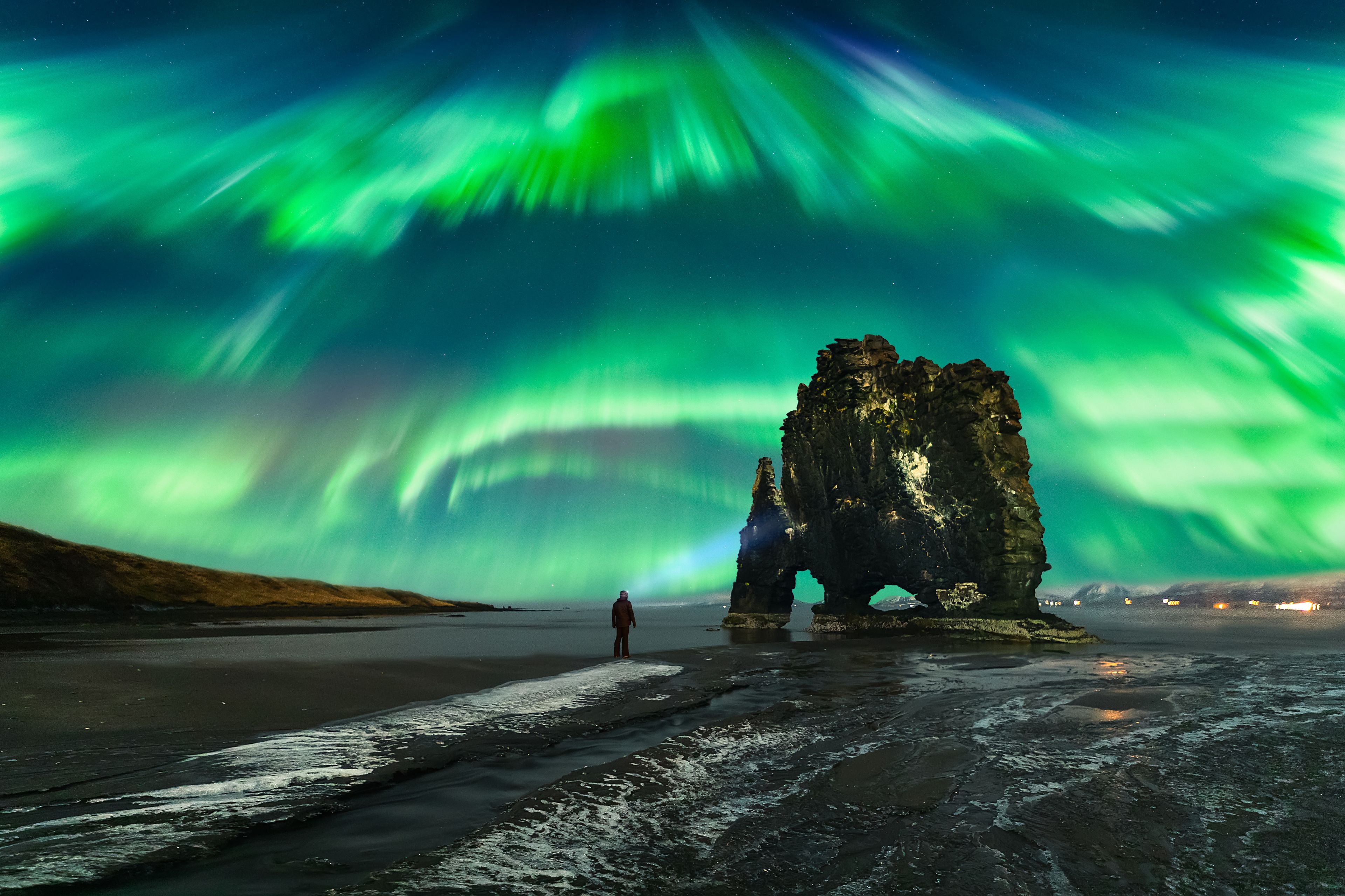 A man with a lantern watches the Northern Light next to the famous volcanic rock formation Hvítserkur.