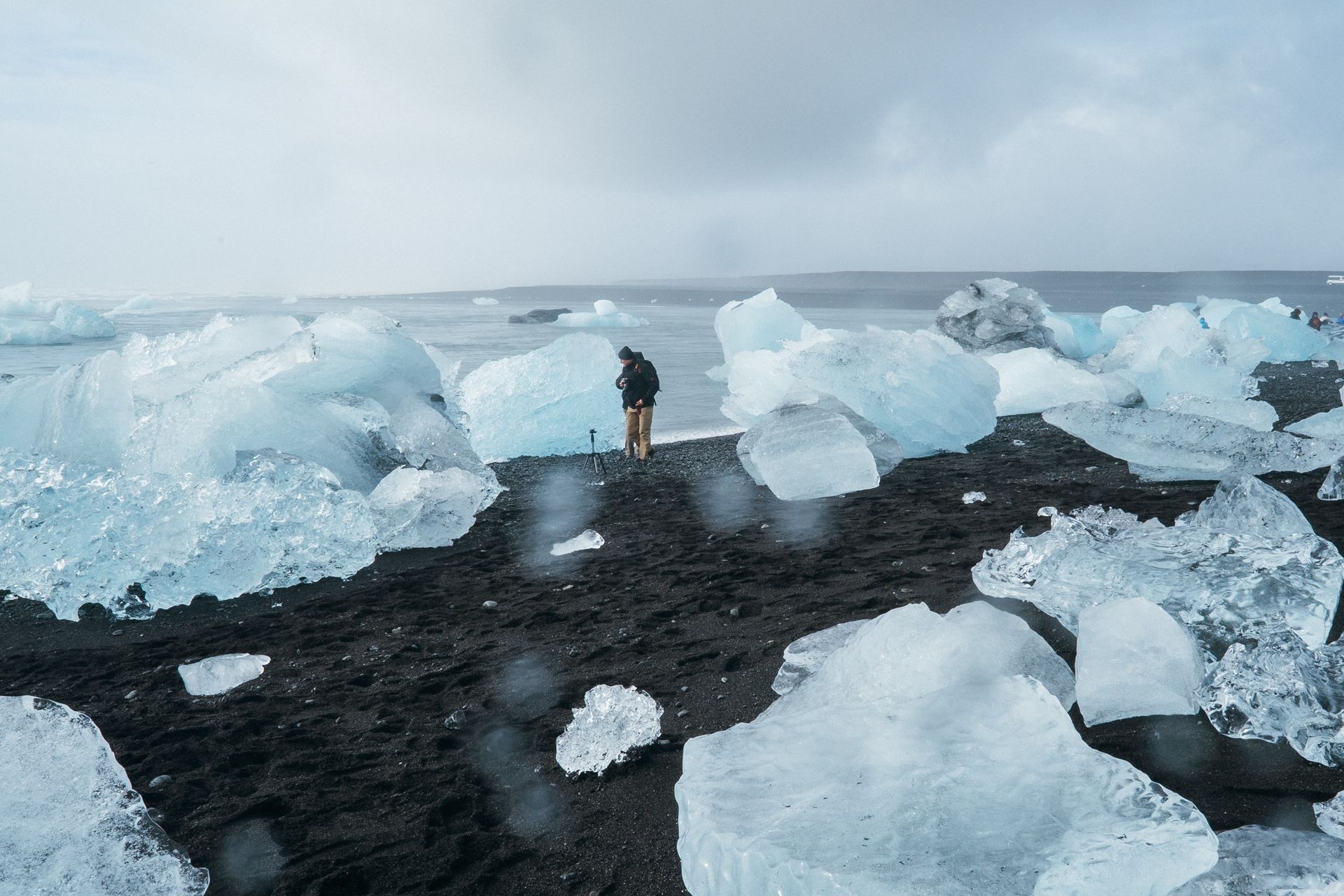 A man walks along the glistening black sand shore of Diamond Beach in Iceland, surrounded by glittering ice chunks scattered along the beach.